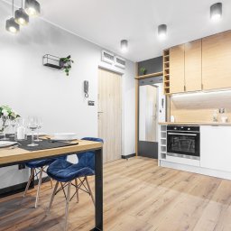 Nowa Perspektywa Meble na wymiar - Jasne wnętrze mieszkania z drewnianą podłogą, drewnianymi blatami kuchennymi i stołem z czarną ramą oraz niebieskimi krzesłami, nakryty do posiłku dla kilku osób.