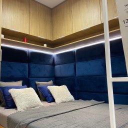 Nowa Perspektywa Meble na wymiar - Nowoczesna sypialnia z zabudową meblową: drewniane szafki pod sufitem i niebieski, tapicerowany zagłówek w formie paneli, oświetlony listwą LED, szara narzuta na łóżku.