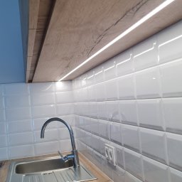 Nowa Perspektywa Meble na wymiar - Nowoczesna kuchnia: blat z drewnopodobnym dekorem, chromowana bateria przy zlewozmywaku i białe, wypukłe płytki na ścianie z oświetleniem LED pod szafkami.