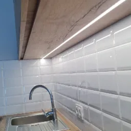 Nowoczesna kuchnia: blat z drewnopodobnym dekorem, chromowana bateria przy zlewozmywaku i białe, wypukłe płytki na ścianie z oświetleniem LED pod szafkami.