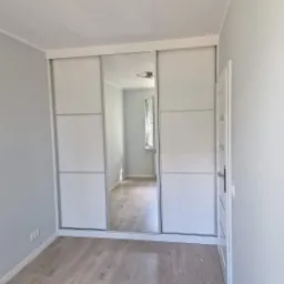 Biała szafa przesuwna z lustrem w minimalistycznej sypialni, widok na puste wnętrze pokoju odbijający się w lustrze, panele podłogowe imitujące drewno.