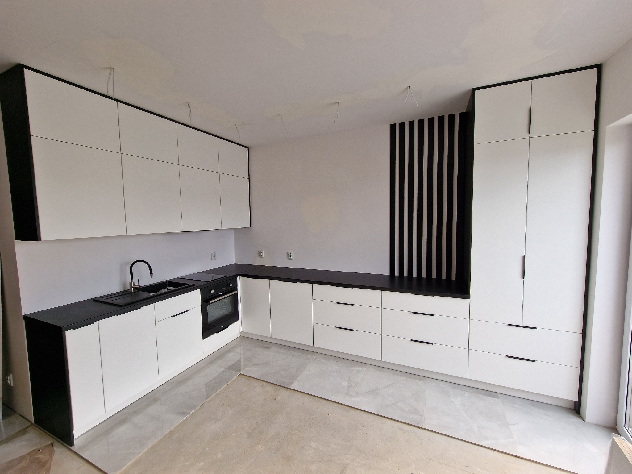 Nowoczesna kuchnia z białymi frontami i czarnymi blatami, widoczne elementy wykończeniowe pod sufitem i fragment podłogi w trakcie układania płytek.