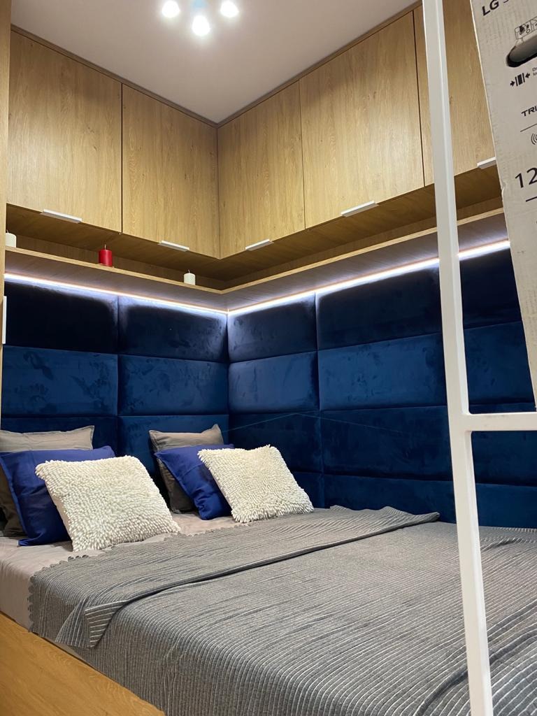 Nowoczesna sypialnia z zabudową meblową: drewniane szafki pod sufitem i niebieski, tapicerowany zagłówek w formie paneli, oświetlony listwą LED, szara narzuta na łóżku.