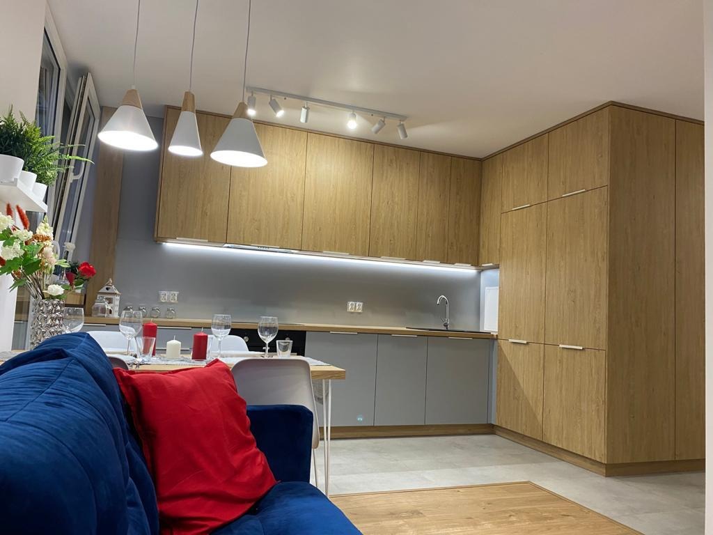 Nowoczesna kuchnia z drewnianymi frontami szafek górnych i szarymi dolnymi, oświetlona wiszącymi lampami i listwą LED pod szafkami, widok z salonu z fragmentem niebieskiej sofy i stołem z nakryciem.