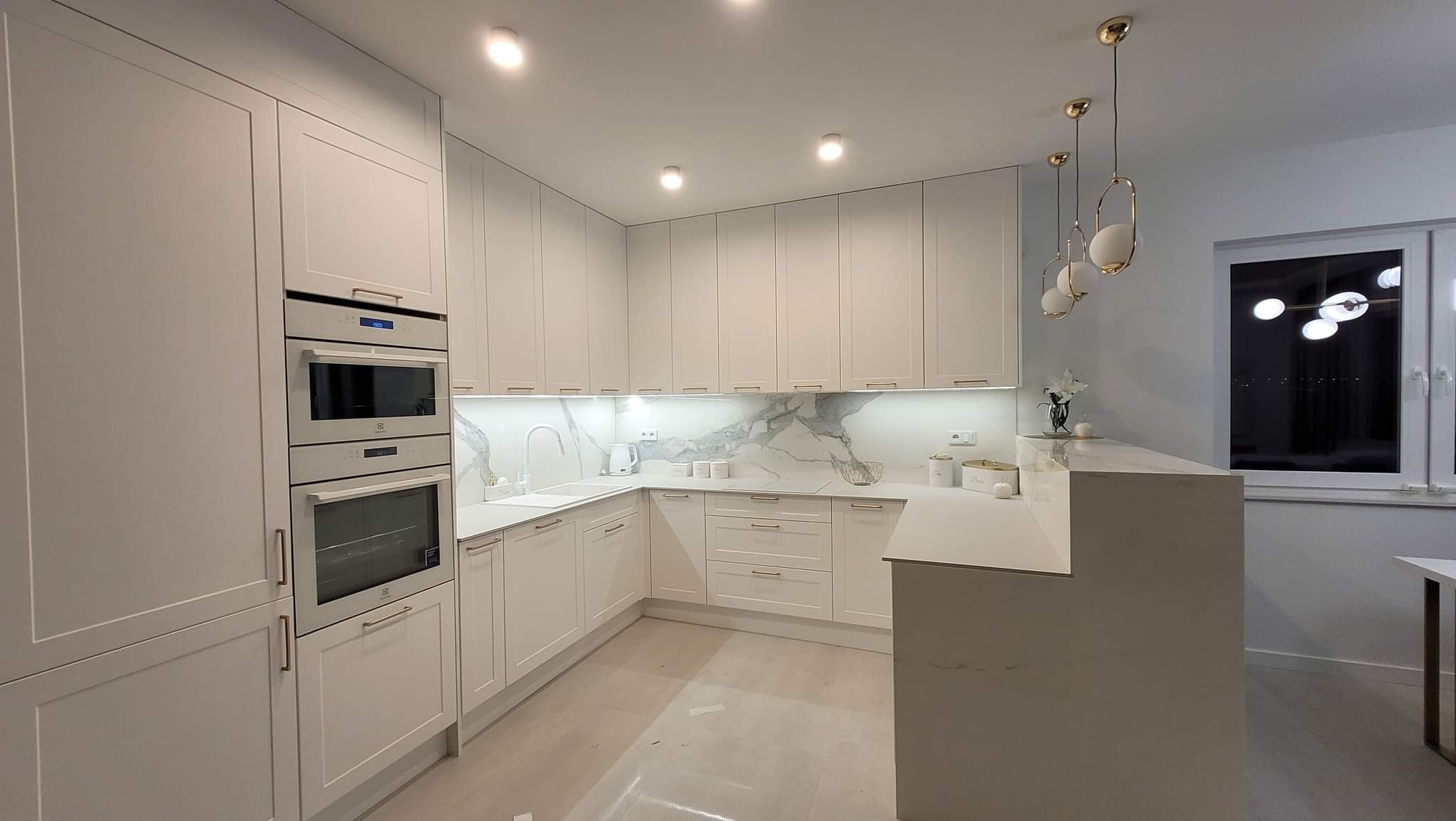Jasna, nowoczesna kuchnia z białymi frontami, marmurowym blatem i złotymi akcentami, oświetlona lampami wiszącymi i listwami LED pod szafkami.