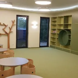 Jasne wnętrze pokoju dziecięcego z drewnianymi dekoracjami w kształcie drzew, okrągłymi stolikami i krzesełkami o minimalistycznym designie oraz zabudową regałową z okrągłymi wnękami.