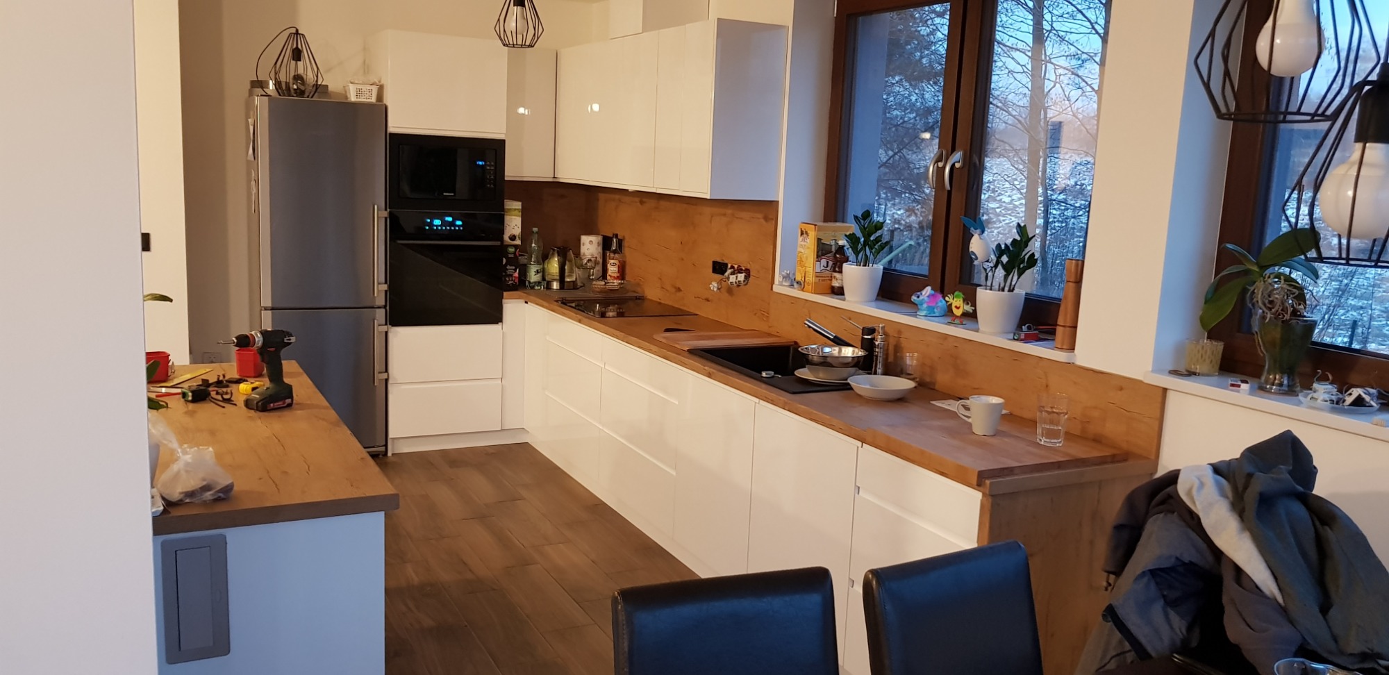 Wnętrze kuchni z białymi szafkami i drewnianym blatem, widoczne okno z zimowym krajobrazem, na blacie kuchennym naczynia i akcesoria, na pierwszym planie fragment stołu z krzesłami.