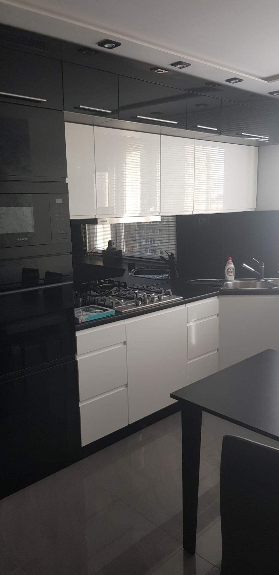 Nowoczesna kuchnia w czarno-białej kolorystyce z błyszczącymi frontami, widok na zabudowę kuchenną z piekarnikiem i płytą gazową, czarny blat i białe szafki dolne, w tle okno z żaluzjami.