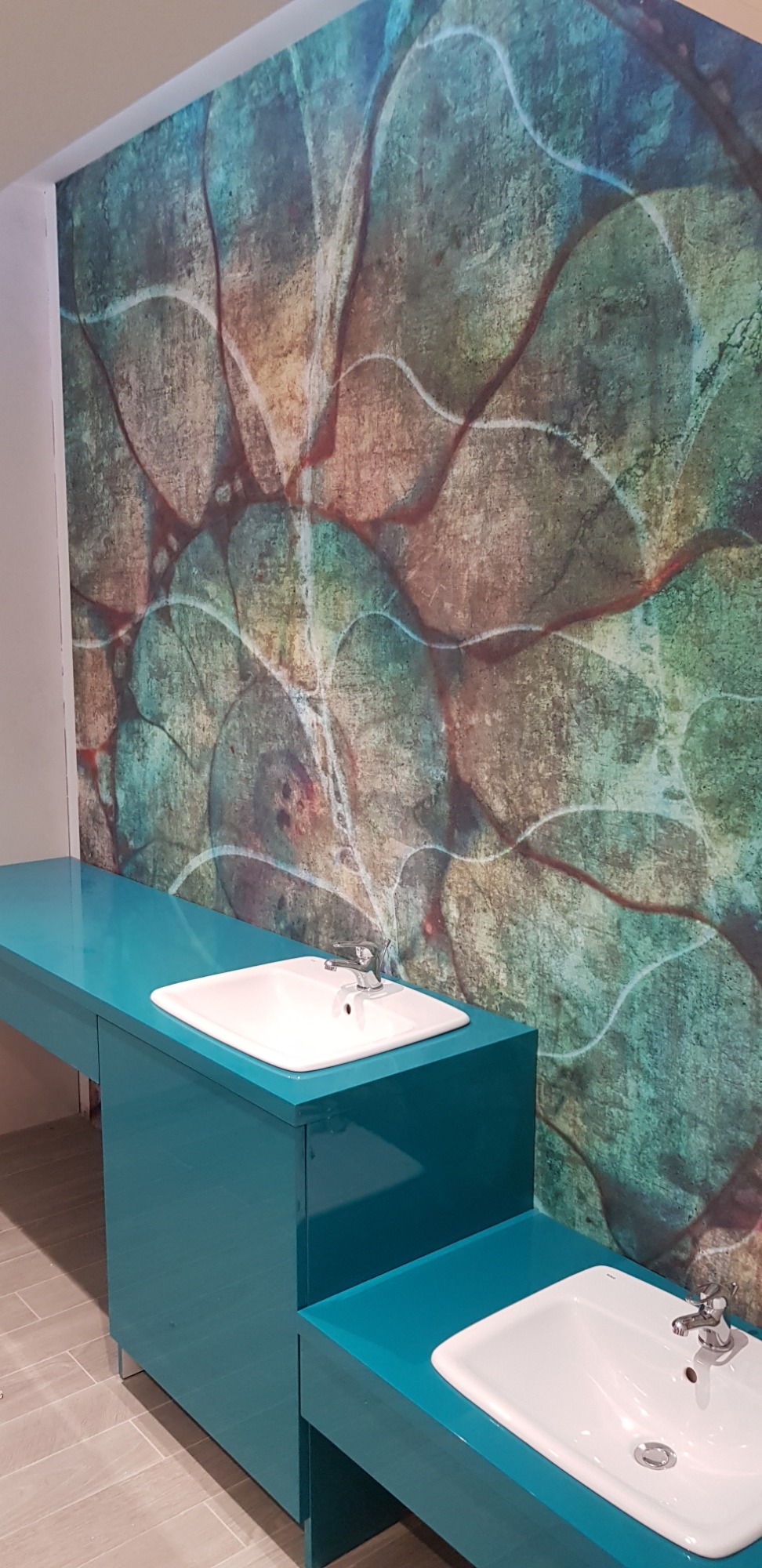 Niebieska szafka łazienkowa z umywalkami na tle ściany z abstrakcyjnym wzorem w odcieniach zieleni, brązu i błękitu.