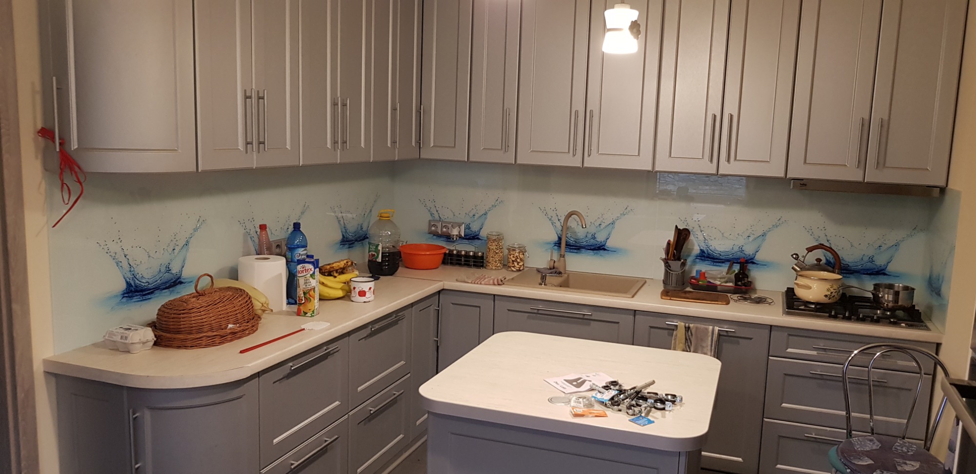 Szara kuchnia na wymiar z grafiką kropli wody na szklanym panelu, widoczne owoce, naczynia i akcesoria kuchenne na blacie.