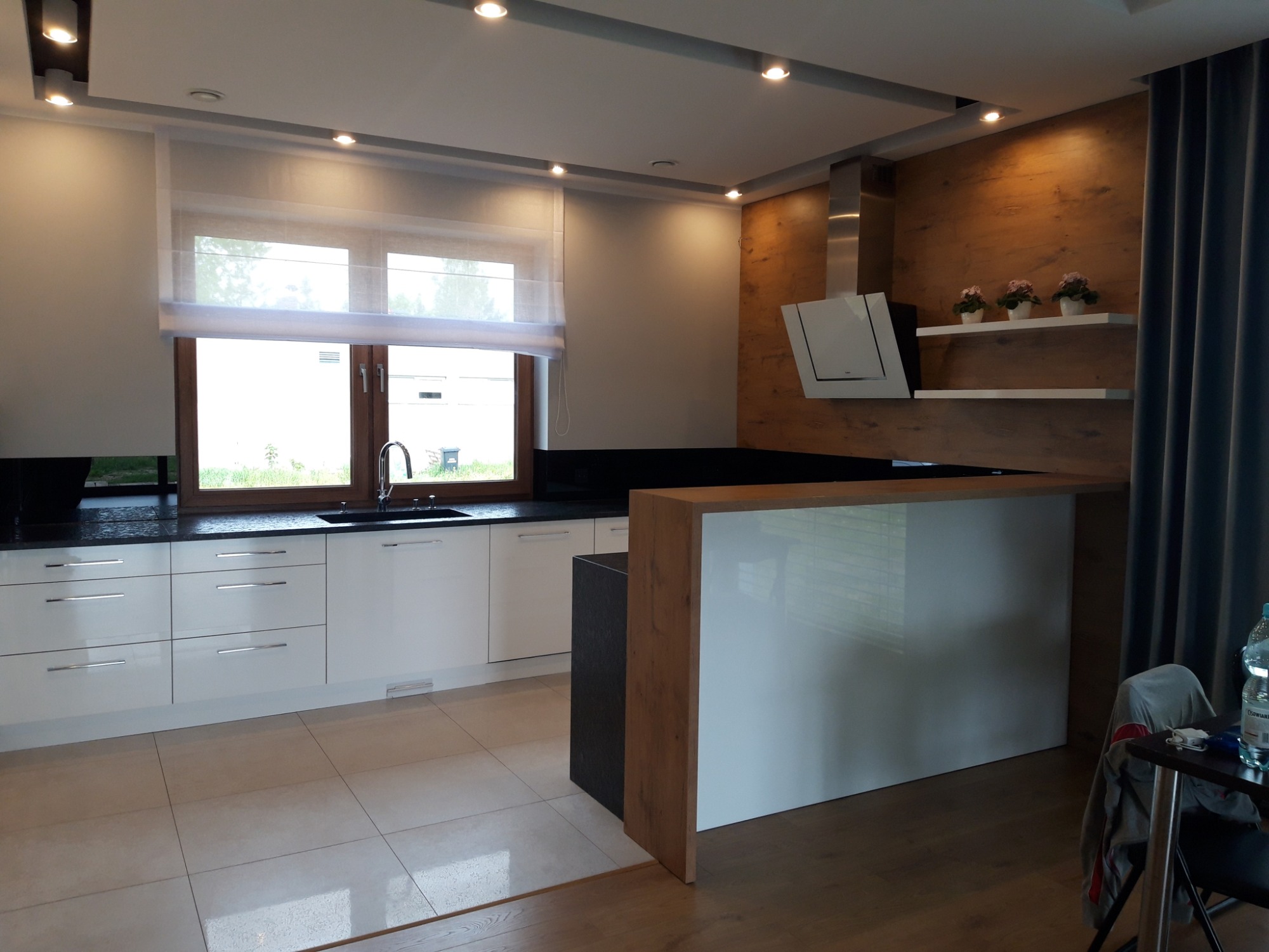 Nowoczesna kuchnia z białymi frontami szafek, czarnym blatem i drewnianą okładziną ściany z okapem oraz półkami z kwiatami, oświetlona punktowymi lampami sufitowymi.