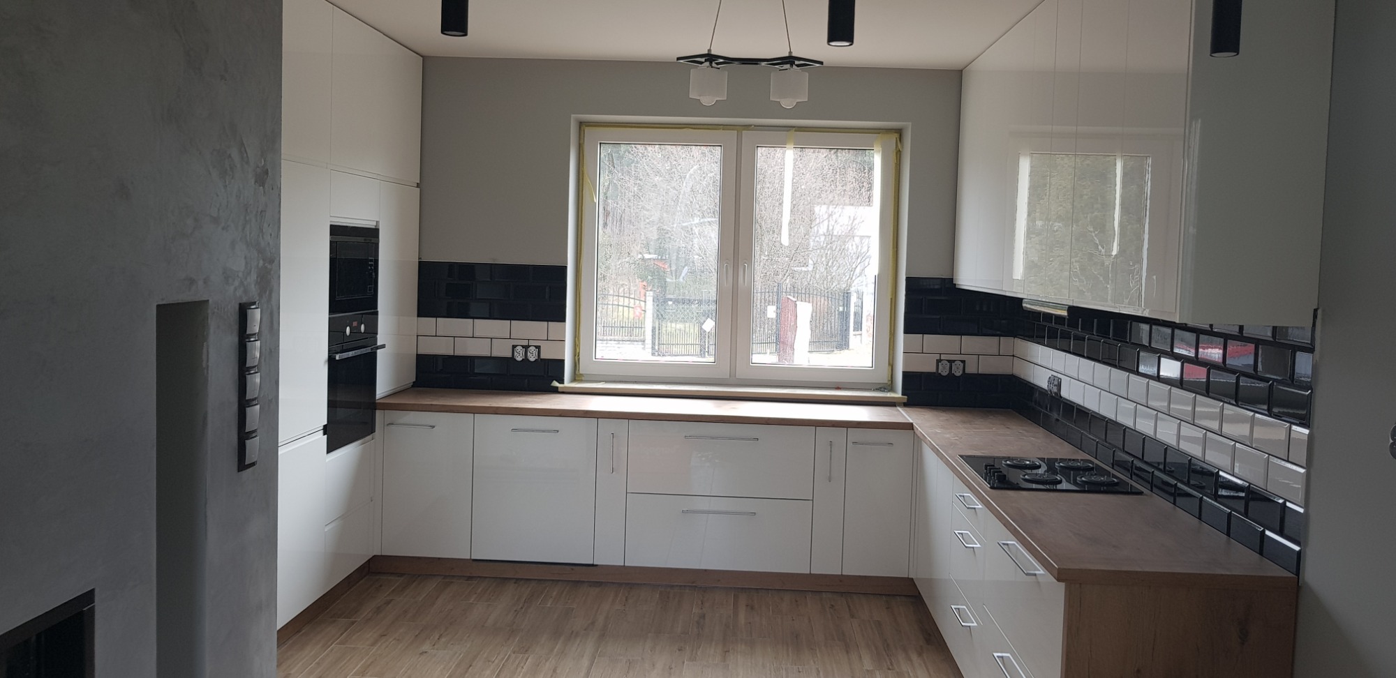 Nowoczesna, jasna kuchnia z białymi, lakierowanymi frontami i drewnianymi blatami w Częstochowie, z czarno-białą glazurą nad blatem i widokiem na zewnątrz przez okno.