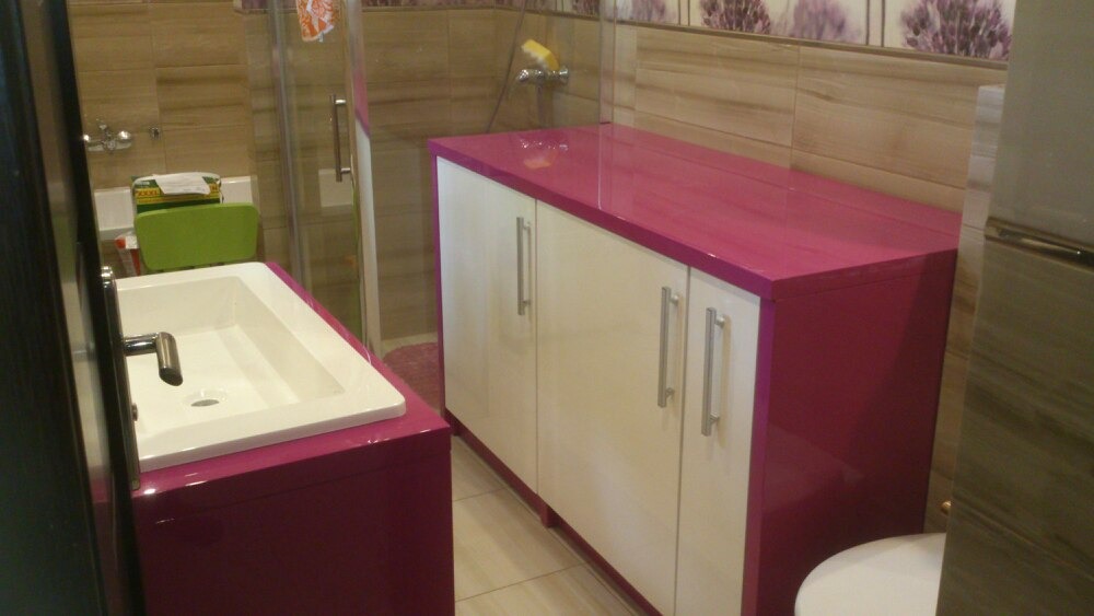 Nowoczesna łazienka z szafką pod umywalkę i szafką stojącą z białymi frontami i różowym blatem, widoczne płytki w kwiaty na ścianie.