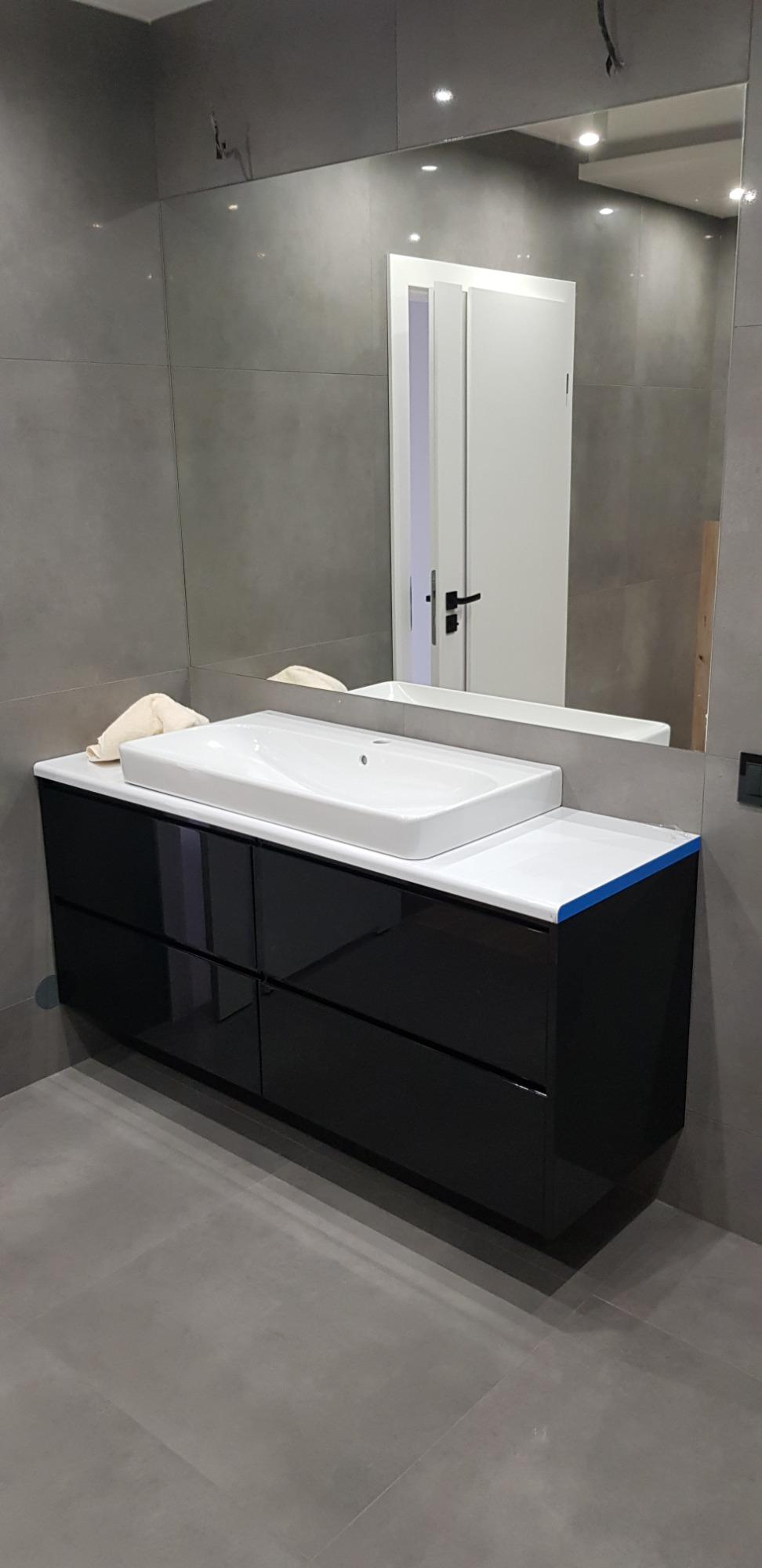 Nowoczesna łazienka z czarną szafką pod umywalką, białym blatem i umywalką oraz dużym lustrem na szarej ścianie z płytkami. Widoczne niedokończone prace wykończeniowe.