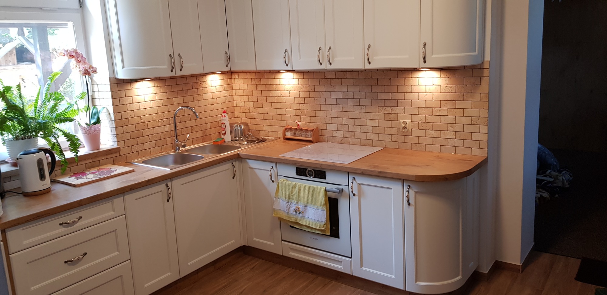Jasna kuchnia z drewnianym blatem, ceglaną mozaiką na ścianie i oświetleniem podszafkowym, z widocznym zlewozmywakiem, płytą indukcyjną i piekarnikiem z zawieszoną ściereczką.