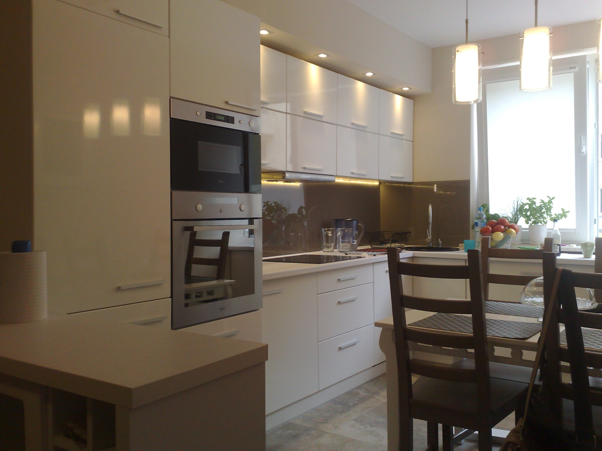 Jasna kuchnia z białymi, lakierowanymi frontami szafek, oświetleniem LED pod szafkami wiszącymi i zabudowanym piekarnikiem z mikrofalówką. Na blacie widoczne szklanki i dzbanek, a przy oknie owoce...