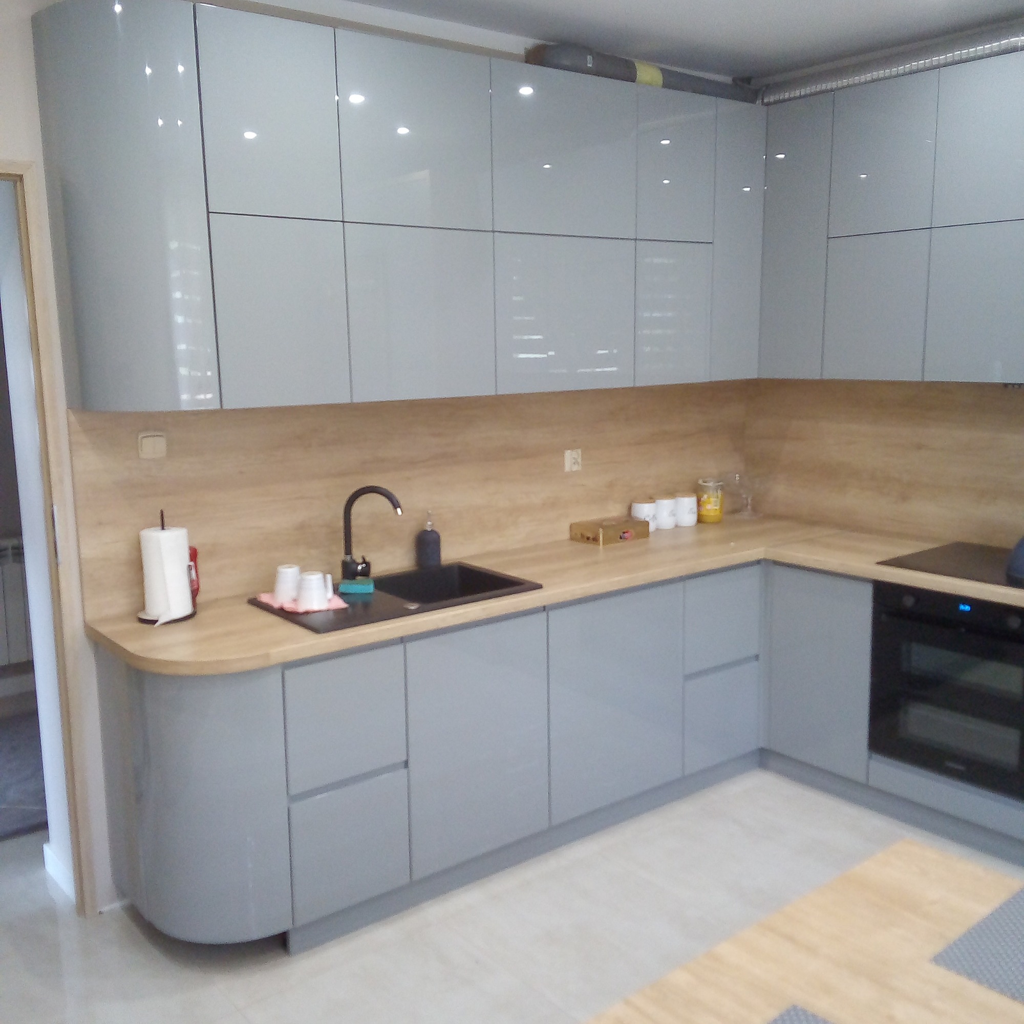 Nowoczesna kuchnia z szarymi, lakierowanymi frontami, drewnianym blatem i czarną armaturą, widok na zabudowę kuchenną ze zlewozmywakiem i płytą indukcyjną.