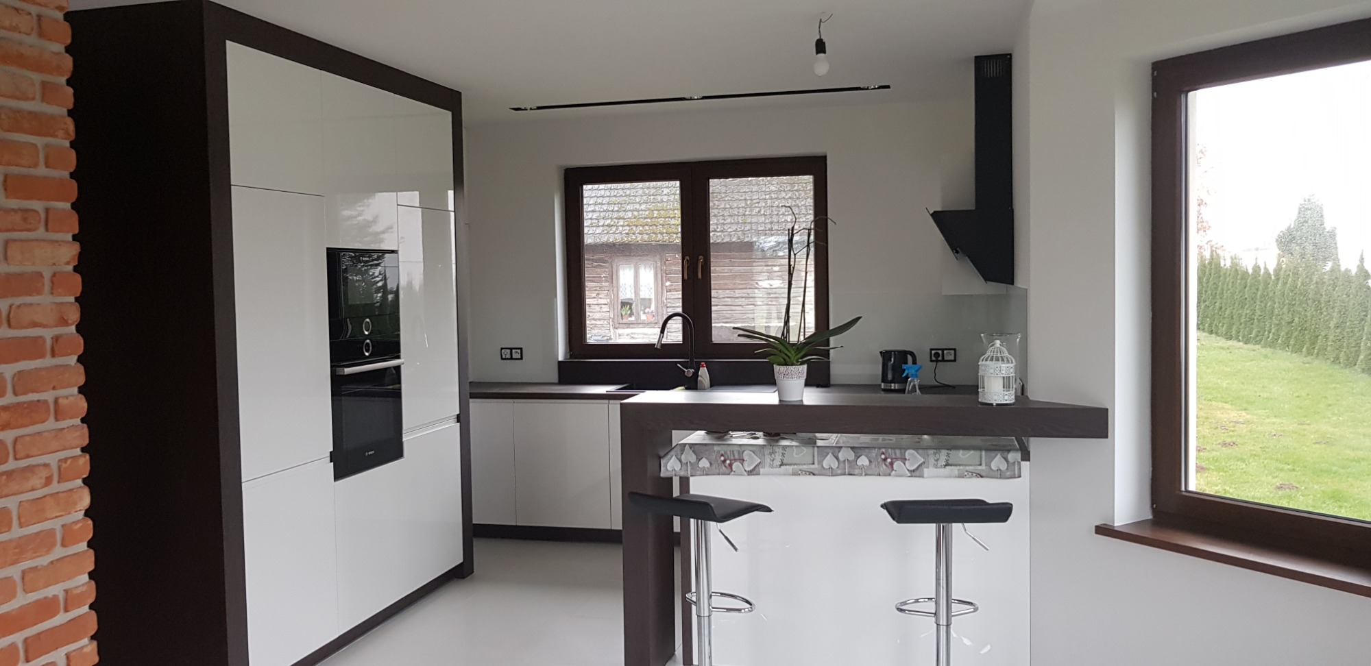 Nowoczesna kuchnia z białymi frontami i ciemnobrązowymi akcentami, wysoka zabudowa z piekarnikiem, wyspa z hokerami, okno z widokiem na ogród. Ciekawy detal: ceglana ściana po lewej stronie.