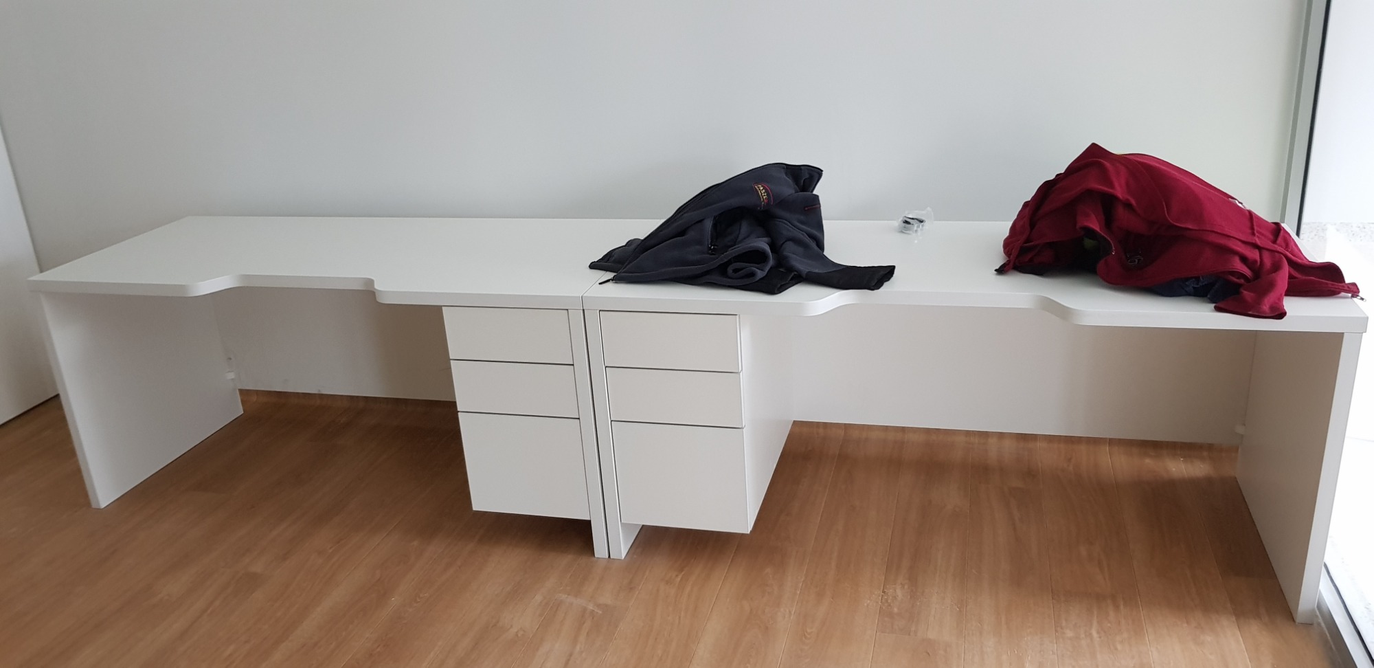 Długi, biały stół zintegrowany z szafkami z szufladami, na którym leżą ubrania, ustawiony na podłodze z paneli w jasnym pomieszczeniu.