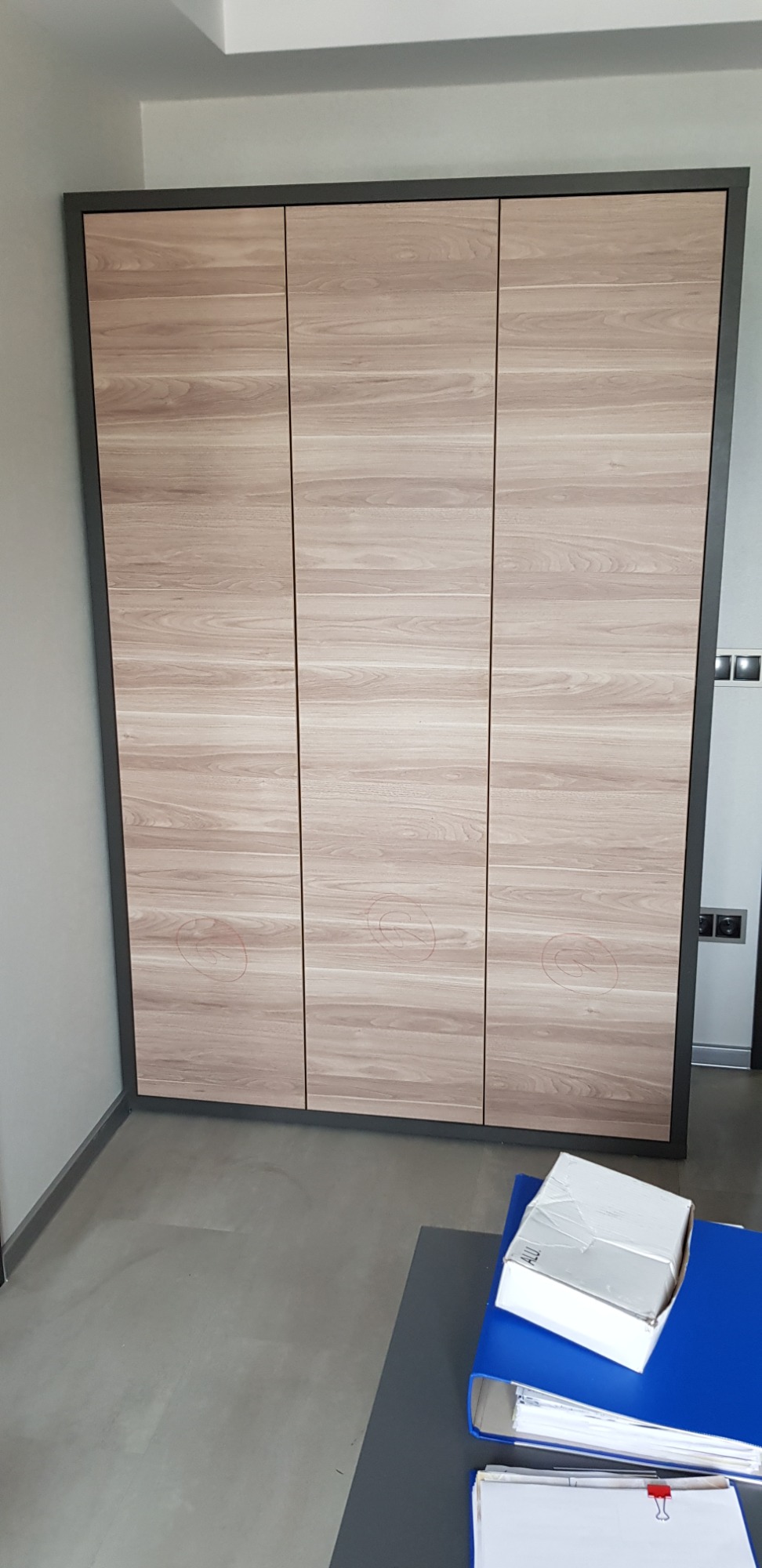 Trzydrzwiowa szafa wnękowa z frontami imitującymi jasne drewno, obramowana ciemnoszarą ramą, z widocznymi lekkimi zabrudzeniami na drzwiach, na tle szarej podłogi i ściany, z elementami biurowymi...