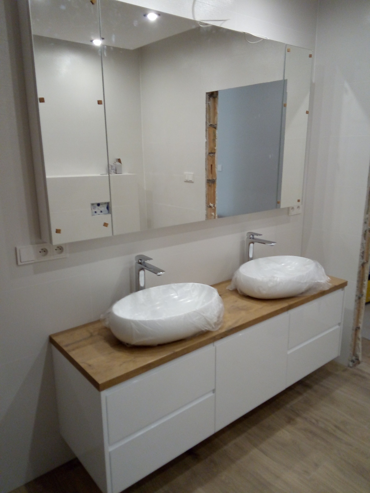 Nowoczesna łazienka w trakcie wykańczania: biała szafka pod umywalki z drewnianym blatem, umywalki owinięte folią ochronną, duże lustro z widocznymi punktami mocowania, surowy stan ścian w tle.