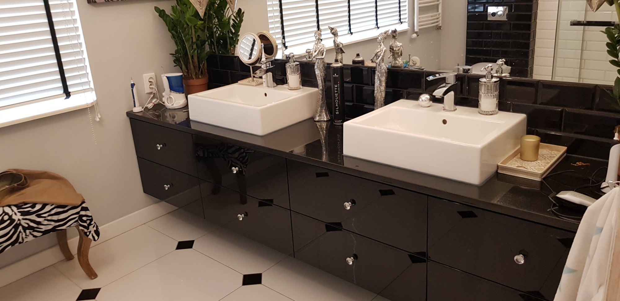 Czarna, lakierowana szafka łazienkowa z dwiema białymi umywalkami nablatowymi, ozdobiona srebrnymi figurkami i akcesoriami, na tle czarnej glazury i białych rolet.