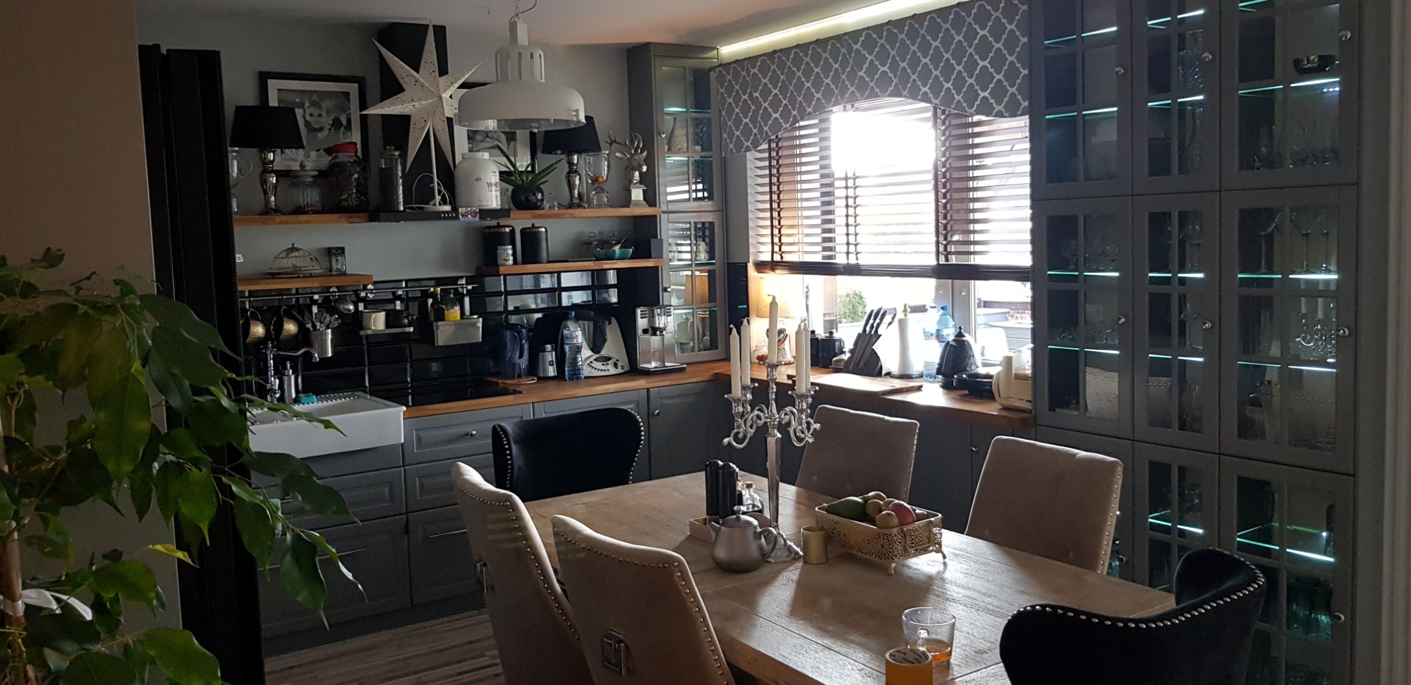 Szara kuchnia z drewnianymi blatami, oświetleniem podszafkowym i przeszklonymi szafkami, widok z perspektywy jadalni z drewnianym stołem i krzesłami.