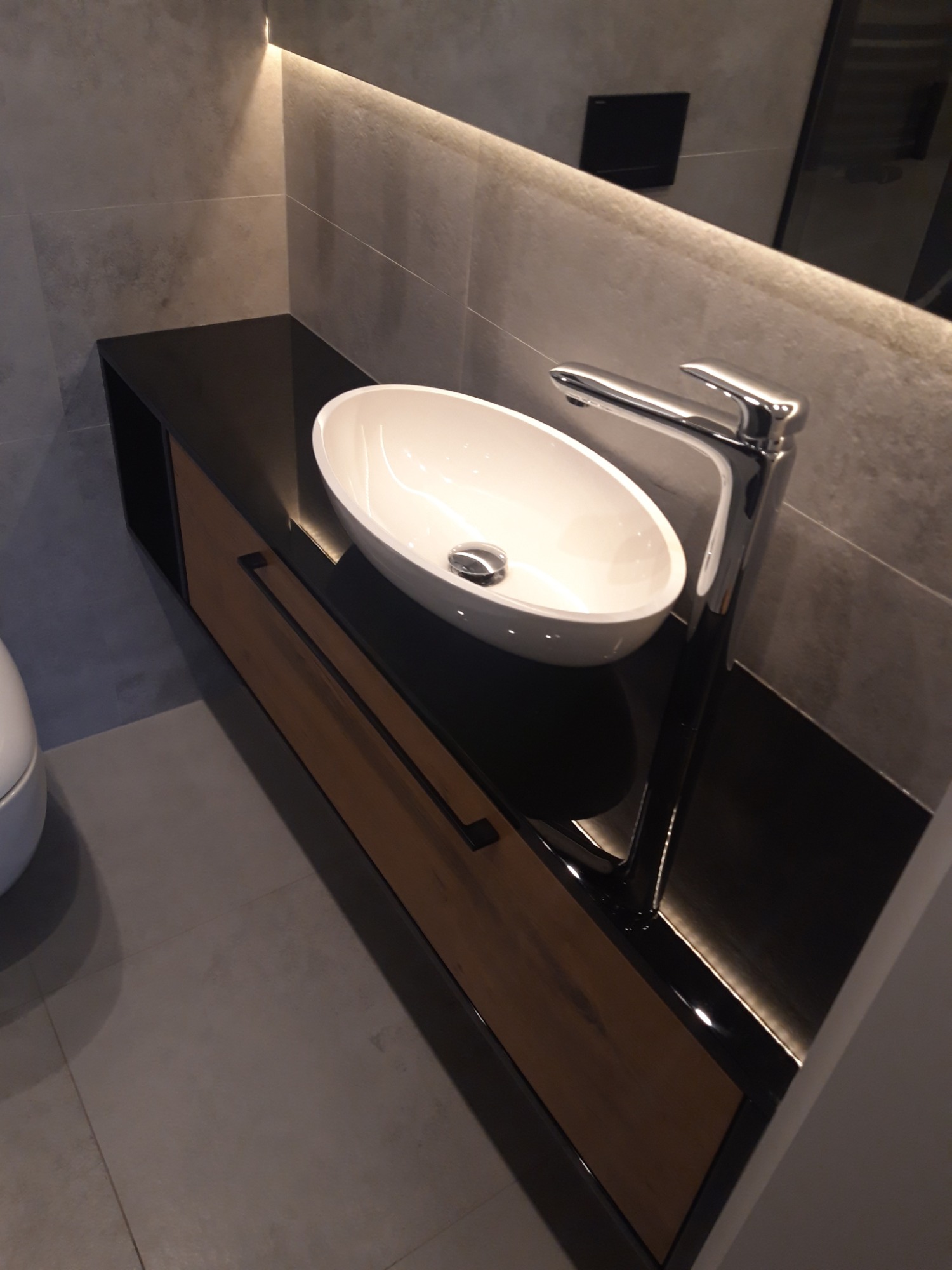 Nowoczesna łazienka z owalną umywalką nablatową, chromowaną baterią i szafką z drewnianym frontem pod czarnym blatem, oświetlona listwą LED.