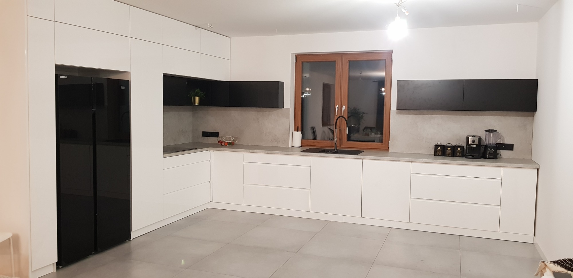 Nowoczesna kuchnia z białymi szafkami dolnymi i czarnymi szafkami górnymi, szarym blatem, czarną lodówką i drewnianym oknem.