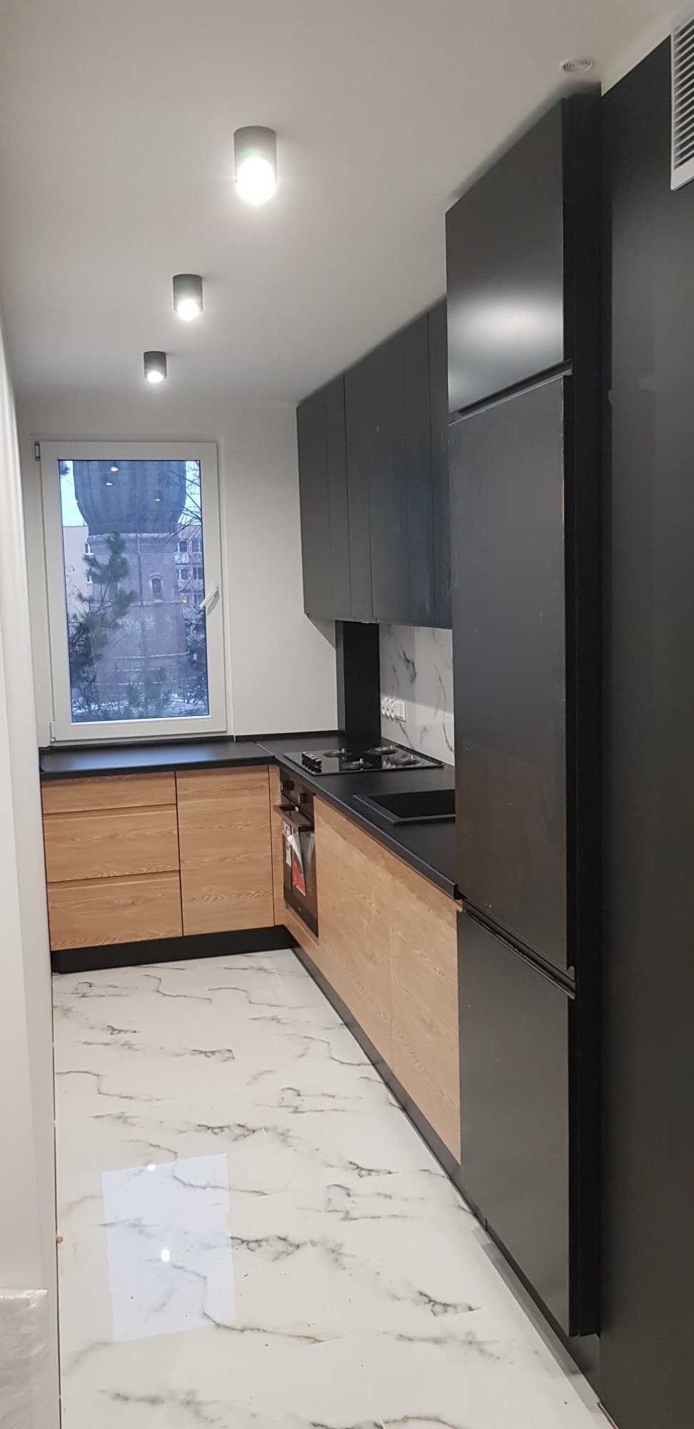 Nowoczesna kuchnia z jasnymi drewnianymi frontami szafek dolnych i czarnymi szafkami górnymi, czarnym blatem oraz marmurową podłogą, oświetlona trzema punktowymi lampami sufitowymi.