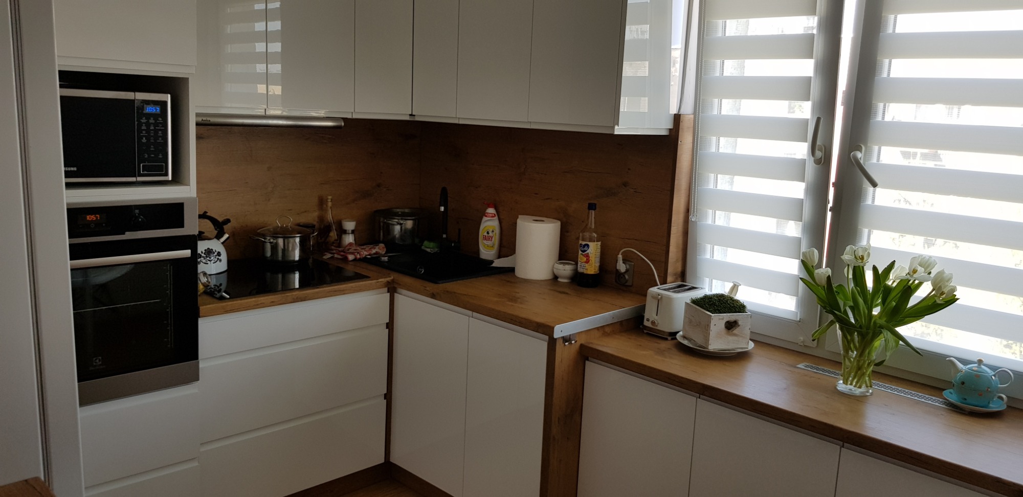 Nowoczesna kuchnia z białymi frontami szafek i drewnianym blatem, wyposażona w piekarnik, mikrofalówkę, płytę indukcyjną i zlewozmywak; na blacie stoją naczynia, przyprawy i kwiaty w wazonie