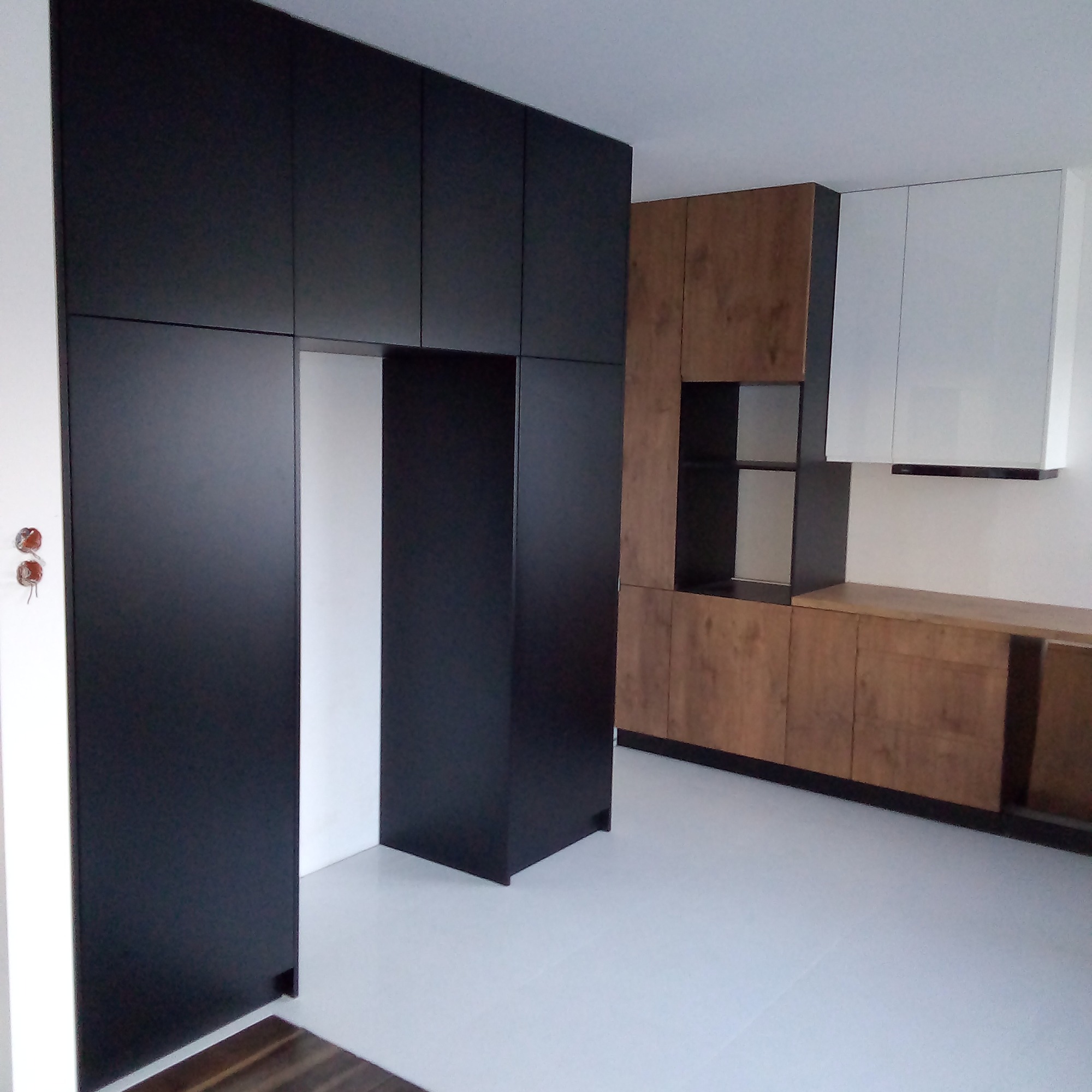 Nowoczesna zabudowa kuchenna w kolorach czerni, drewna i bieli, z minimalistycznymi frontami i ukrytymi szafkami, na tle jasnej podłogi.