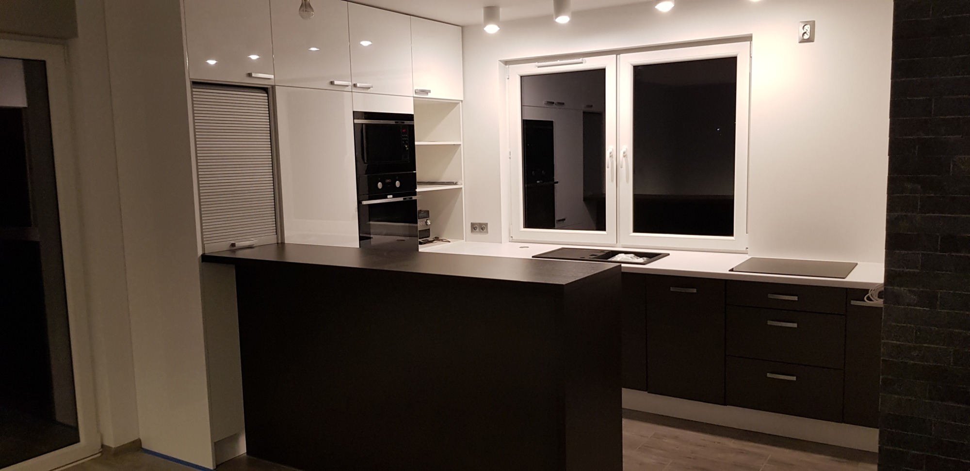 Nowoczesna kuchnia z białymi szafkami górnymi, czarnymi szafkami dolnymi i blatem w kolorze ciemnego drewna, widoczna zabudowa piekarnika i płyty grzewczej, oświetlenie punktowe.