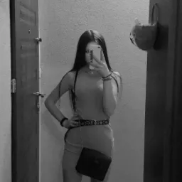 Czarno-białe selfie w lustrze: młoda kobieta w dopasowanej sukience, z torebką na łańcuszku i paskiem z okrągłymi elementami, pozuje w przedpokoju przed otwartymi drzwiami.