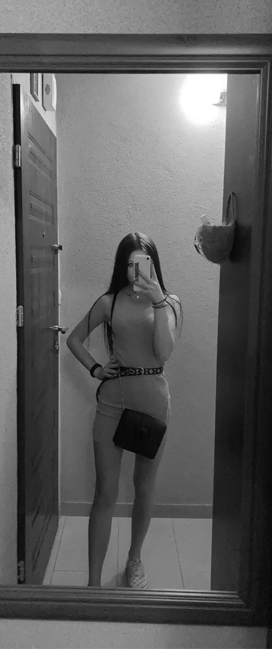 Czarno-białe selfie w lustrze: młoda kobieta w dopasowanej sukience, z torebką na łańcuszku i paskiem z okrągłymi elementami, pozuje w przedpokoju przed otwartymi drzwiami.