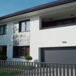 Nowoczesny dom jednorodzinny z białymi ścianami, szarą elewacją z kamienia, balkonem i szarą bramą garażową, widziany zza metalowego ogrodzenia w słoneczny dzień. Logo firmy Unibud w prawym g&oacute;rnym...