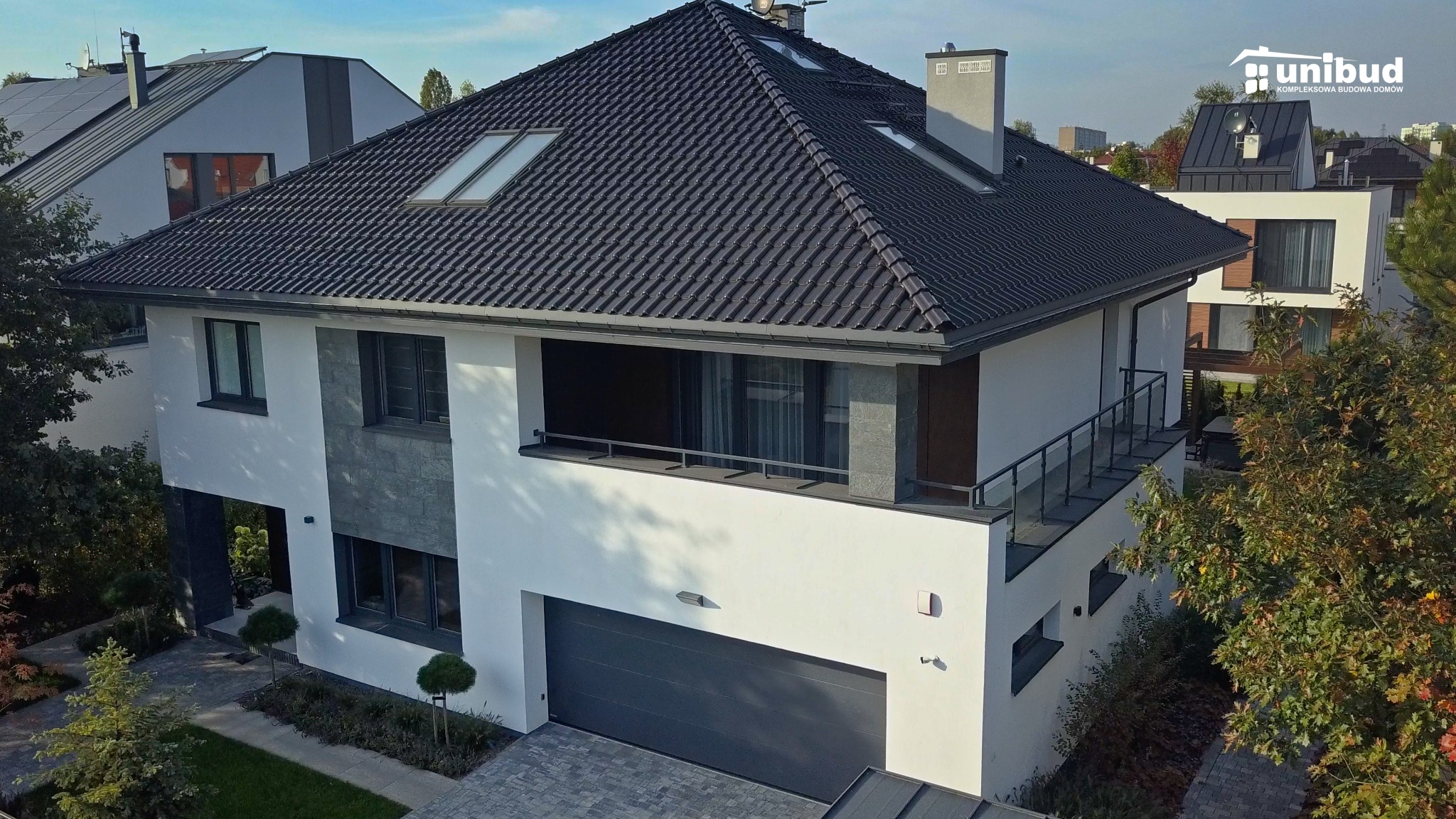 Nowoczesny dwupiętrowy dom z grafitową dachówką, balkonem z barierką i garażem, widok z góry, z logo firmy budowlanej w prawym górnym rogu.
