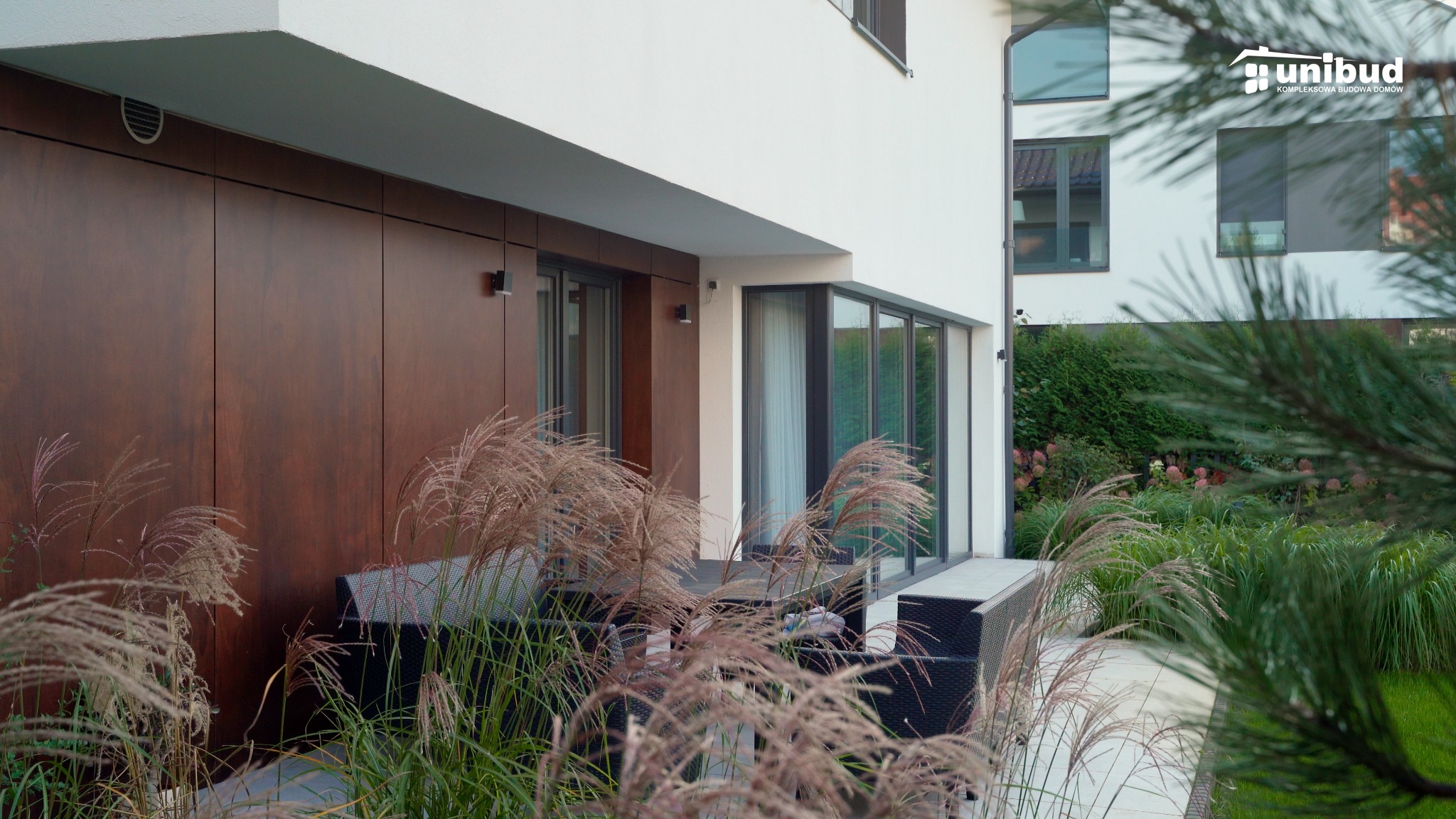 Nowoczesny dom murowany z drewnianą elewacją i dużymi oknami tarasowymi, widok z ogrodu z ozdobnymi trawami, logo firmy Unibud w prawym górnym rogu.