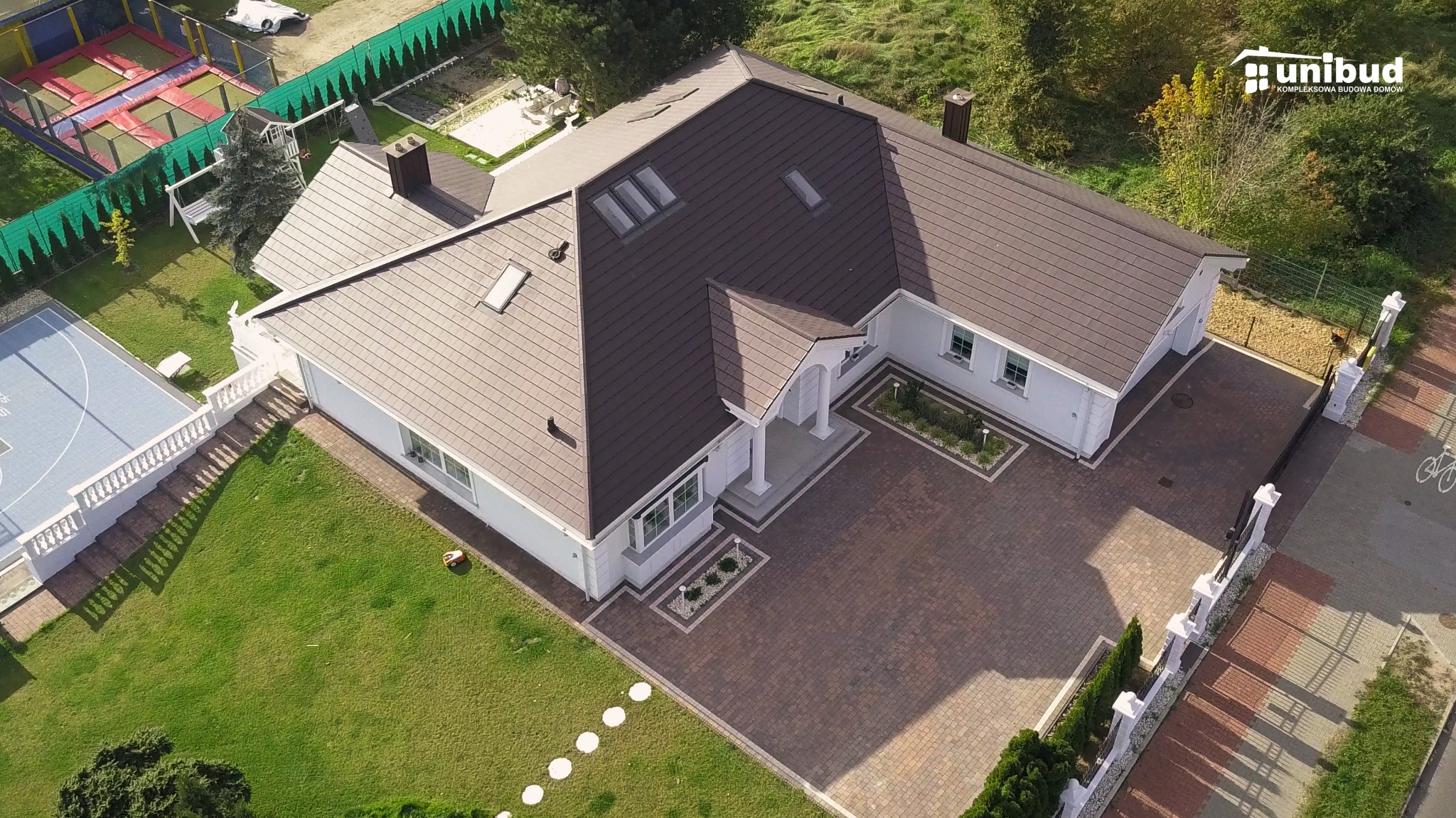 Widok z lotu ptaka na nowoczesny, biały dom z brązowym dachem, starannie zagospodarowanym terenem, w tym trawnikiem, kostką brukową i boiskiem do koszykówki, z logo firmy budowlanej w prawym górnym...