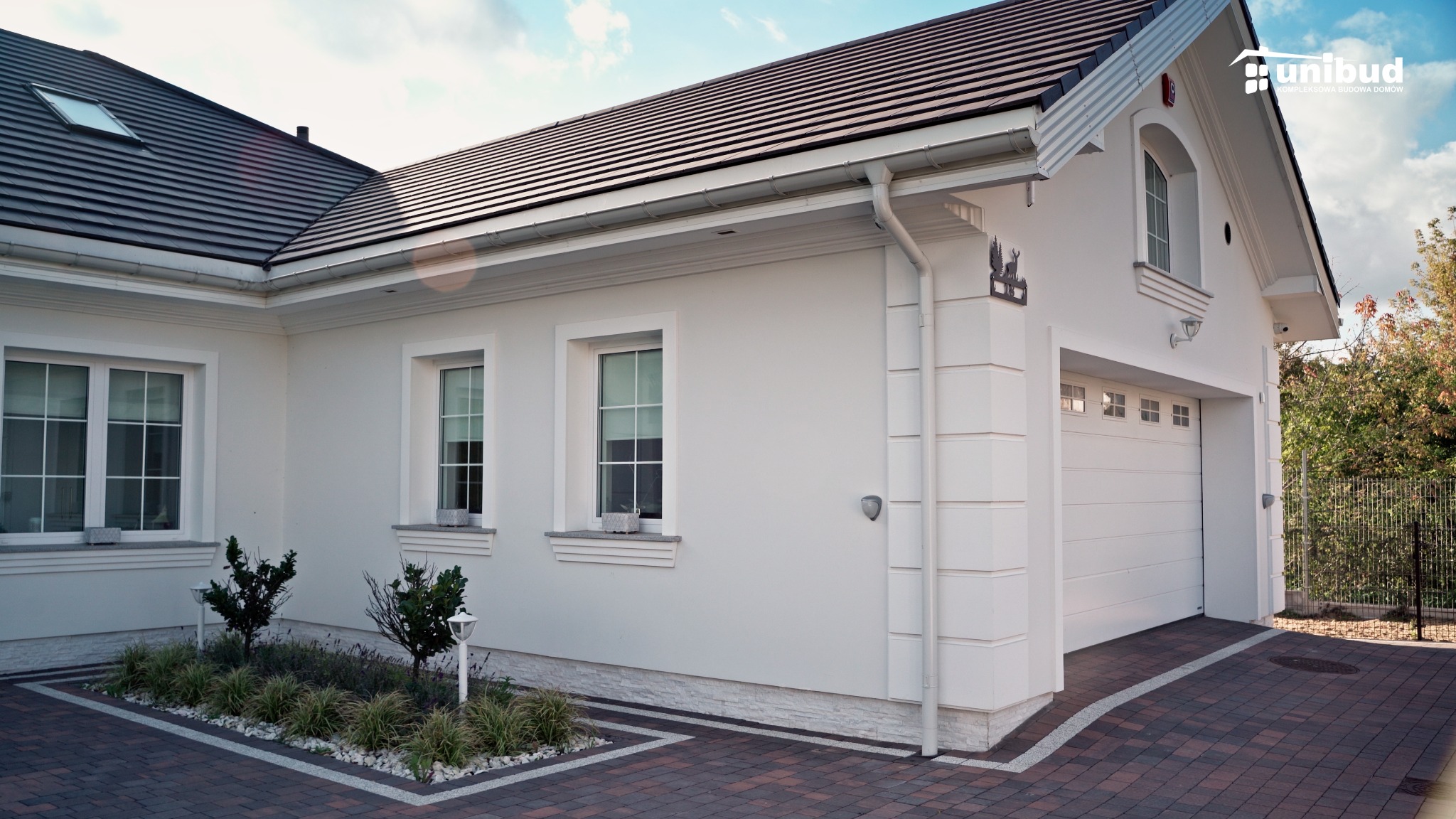 Nowoczesny, jednopiętrowy dom z białymi ścianami, ciemną dachówką i garażem, otoczony kostką brukową i niską zielenią, z logo firmy Unibud w prawym górnym rogu.
