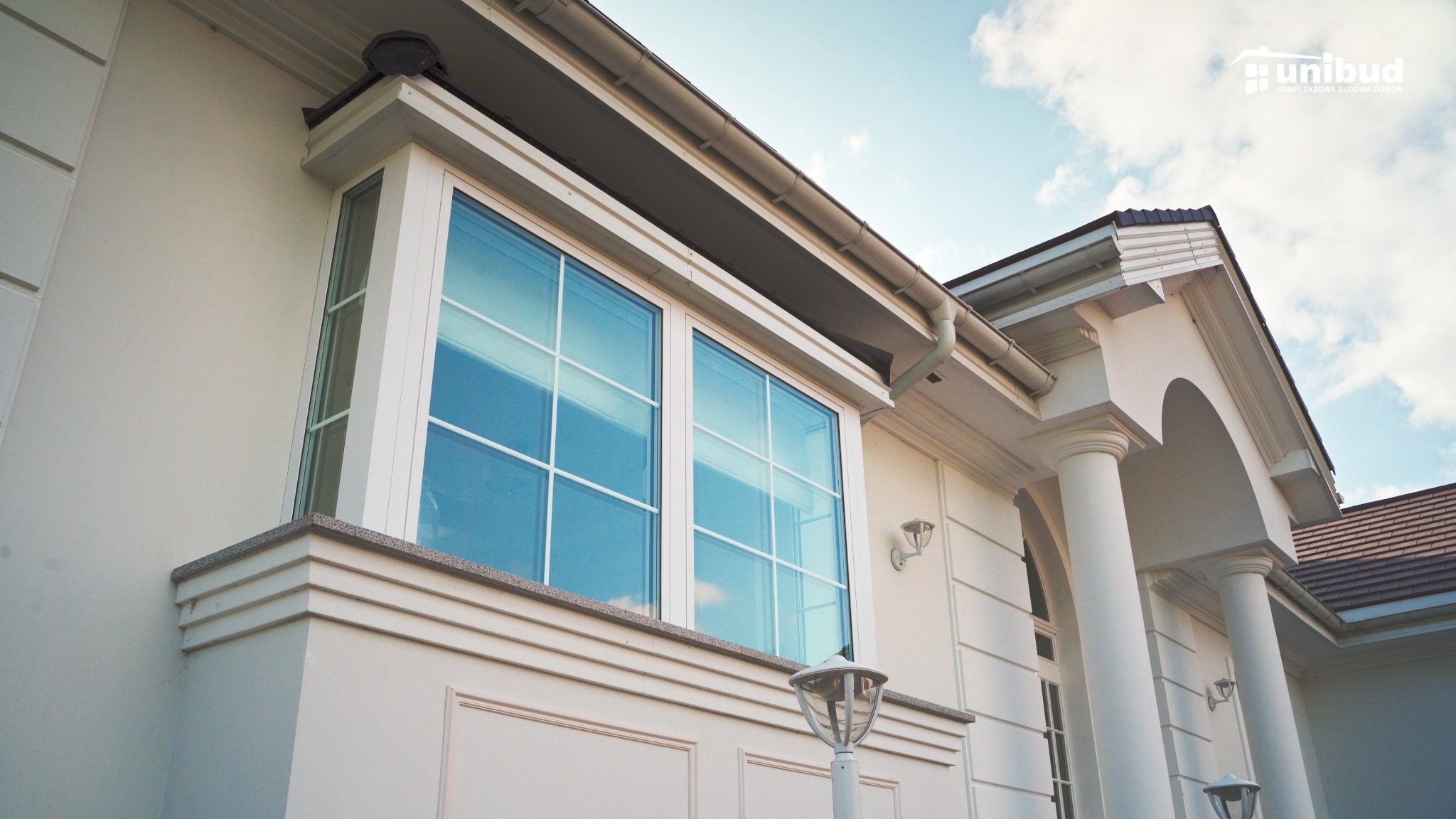 Elewacja domu murowanego z dużym oknem wykuszowym i kolumnami przy wejściu, widoczne detale architektoniczne i logo firmy budowlanej w prawym górnym rogu.