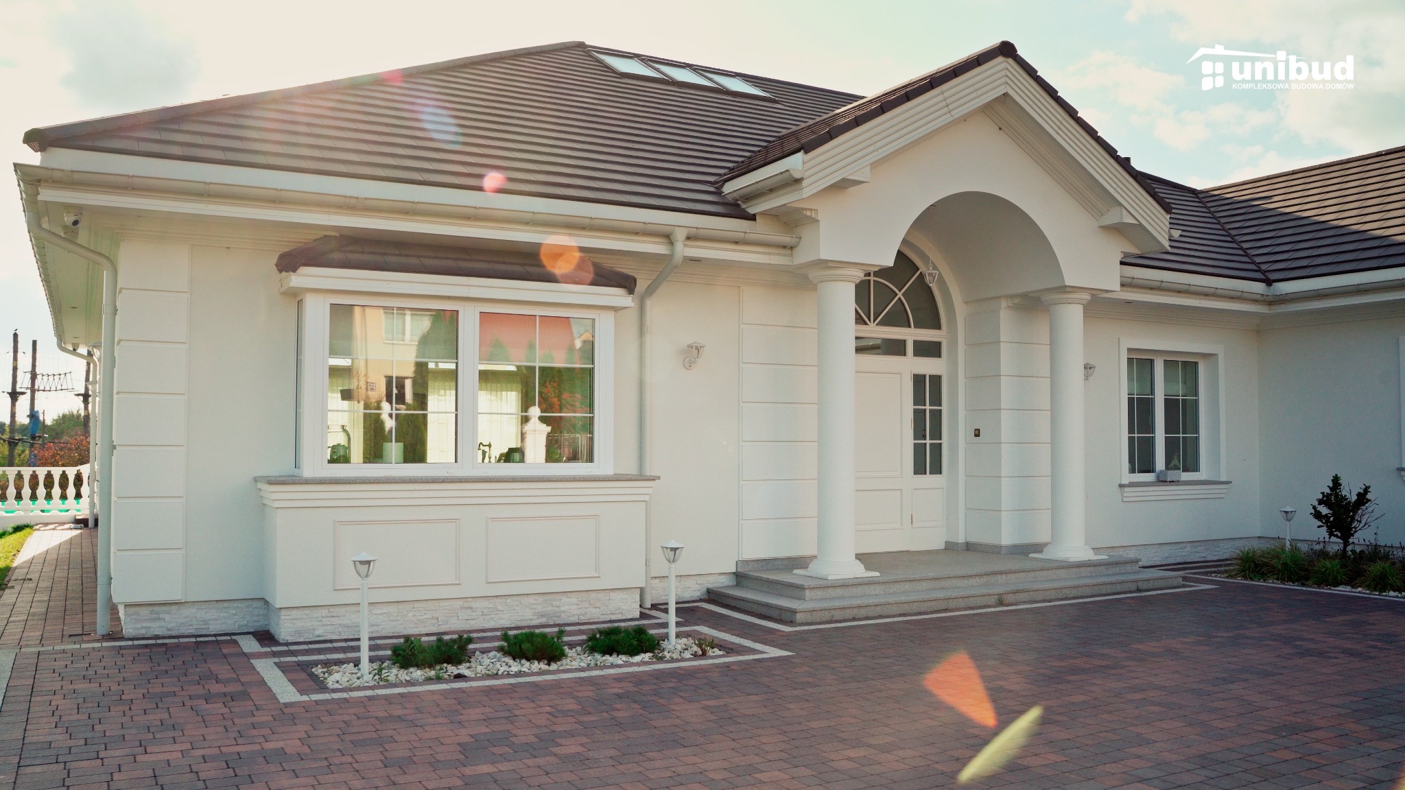 Elegancki, parterowy dom z białymi ścianami, brązowym dachem i kostką brukową, z kolumnami przy wejściu i oknem wykuszowym, oświetlony promieniami słońca.