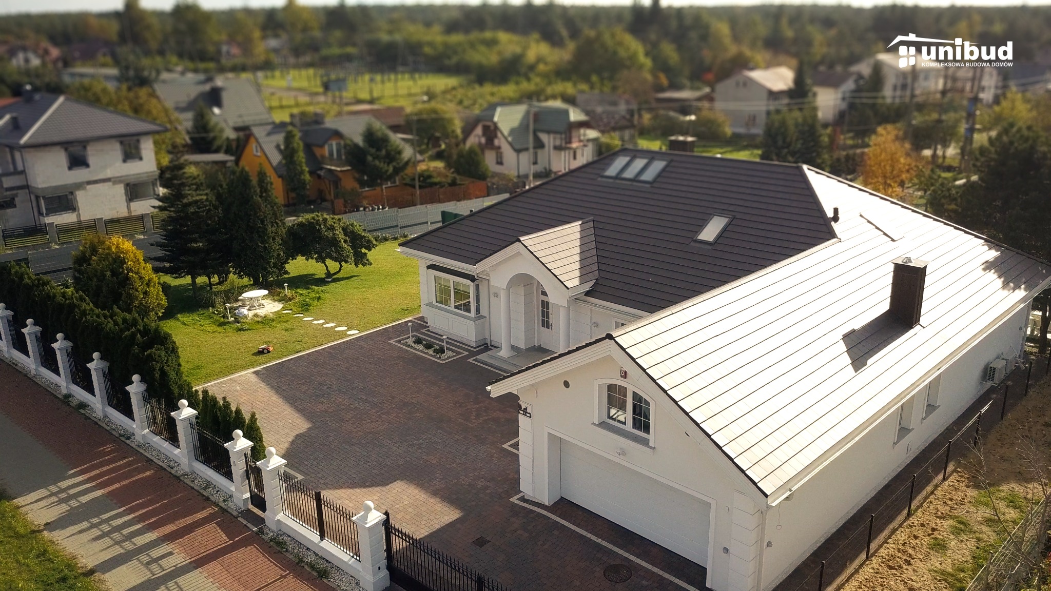 Widok z góry na elegancki, parterowy dom z garażem, białą elewacją i ciemną dachówką, otoczony zadbanym ogrodem i brukowanym podjazdem, widziany z perspektywy drona.