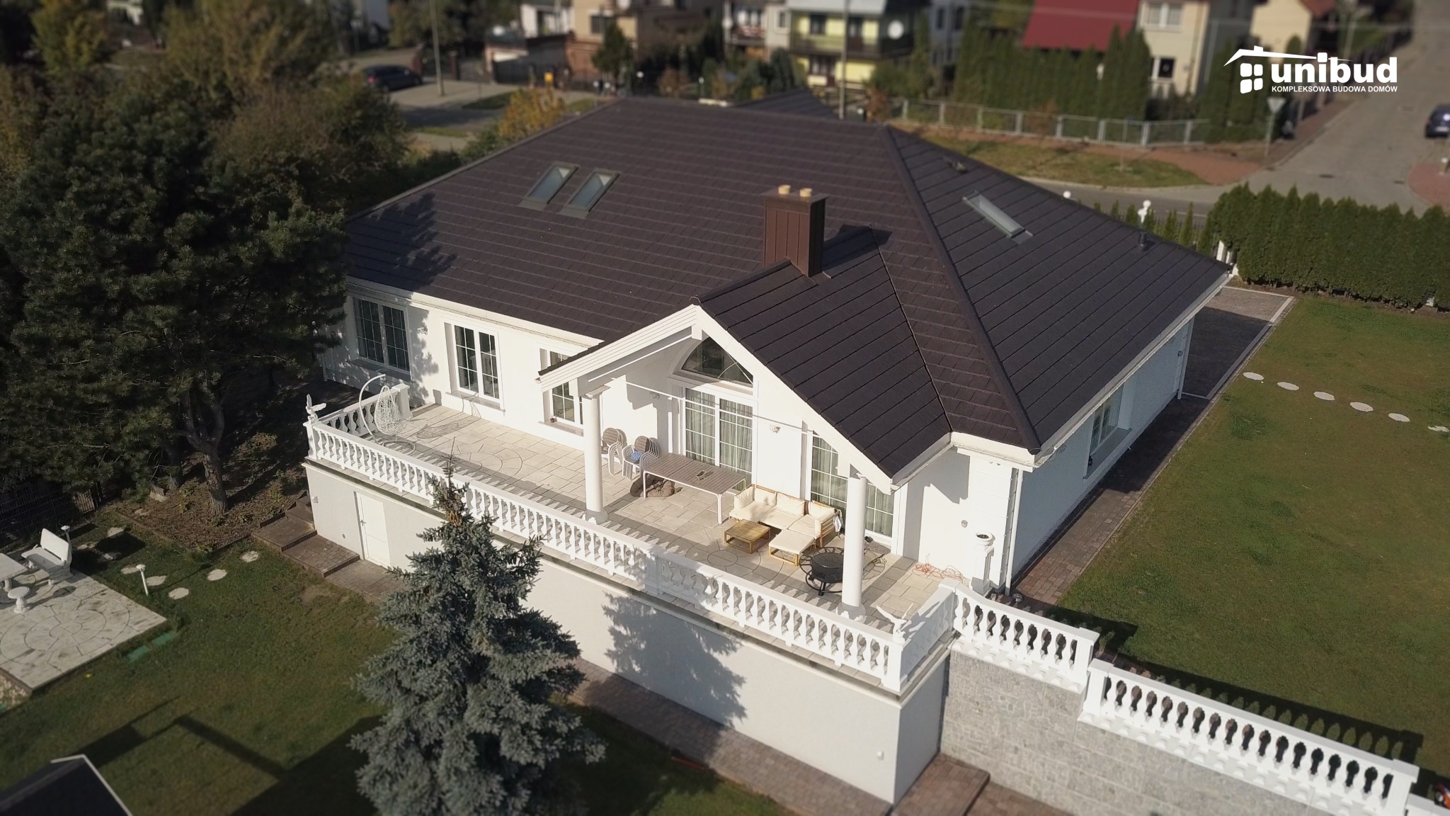 Nowoczesny, parterowy dom z ciemnobrązowym dachem, białymi ścianami i przestronnym tarasem z białymi balustradami, widok z lotu ptaka.