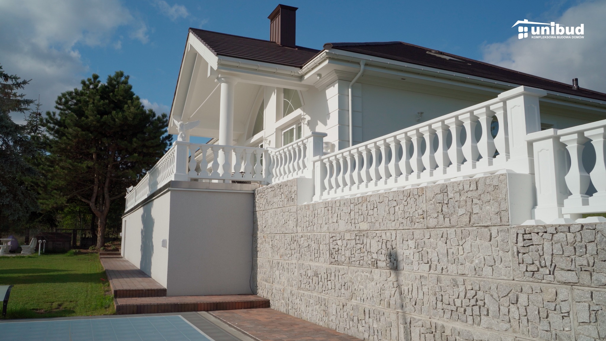 Elewacja domu z białymi kolumnami i ozdobną balustradą, wykończona kamieniem elewacyjnym w odcieniach szarości, z widocznym logo firmy budowlanej w prawym górnym rogu.
