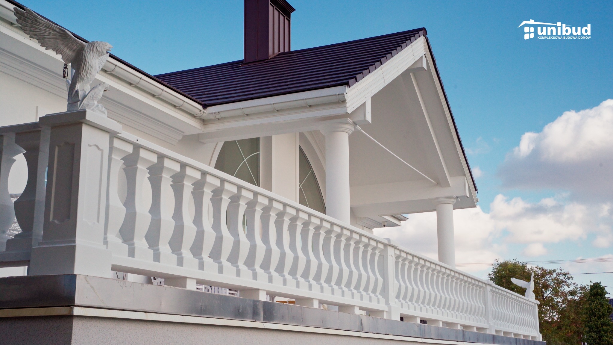 Biały dom murowany z balkonem ozdobionym balustradą i rzeźbą orła, widoczne brązowe pokrycie dachowe i logo firmy budowlanej Unibud.