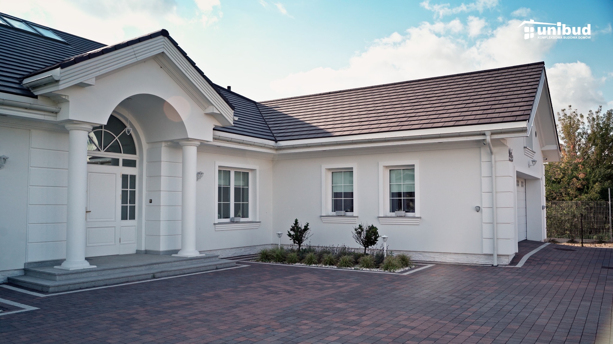 Elegancki, parterowy dom z białymi ścianami, ciemną dachówką i kolumnami przy wejściu, wyłożony kostką brukową w odcieniach czerwieni i brązu, z logo firmy budowlanej w prawym górnym rogu.