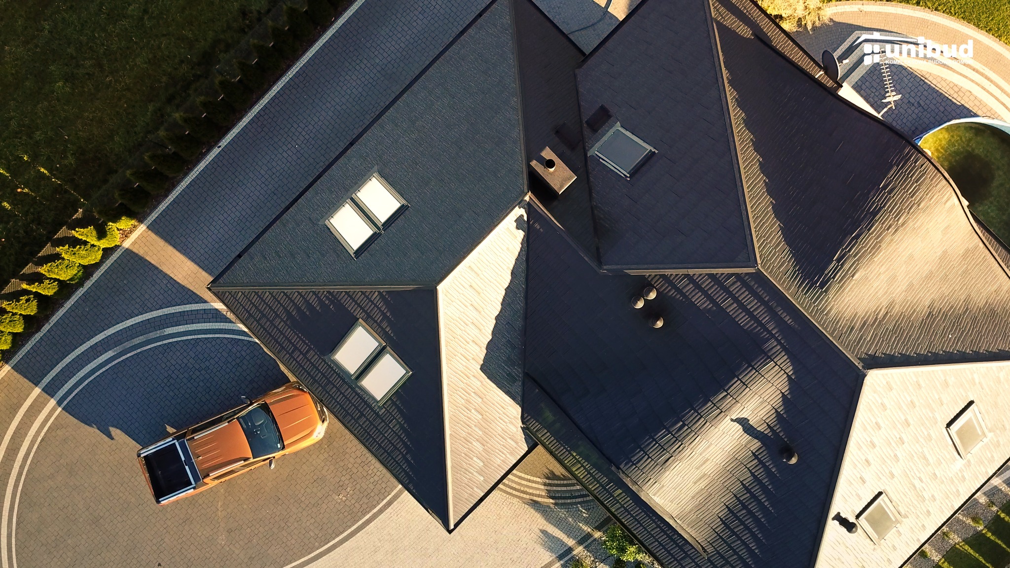 Widok z lotu ptaka na nowoczesny dom z ciemnym dachem, lukarnami i pomarańczowym pickupem zaparkowanym na brukowanym podjeździe.