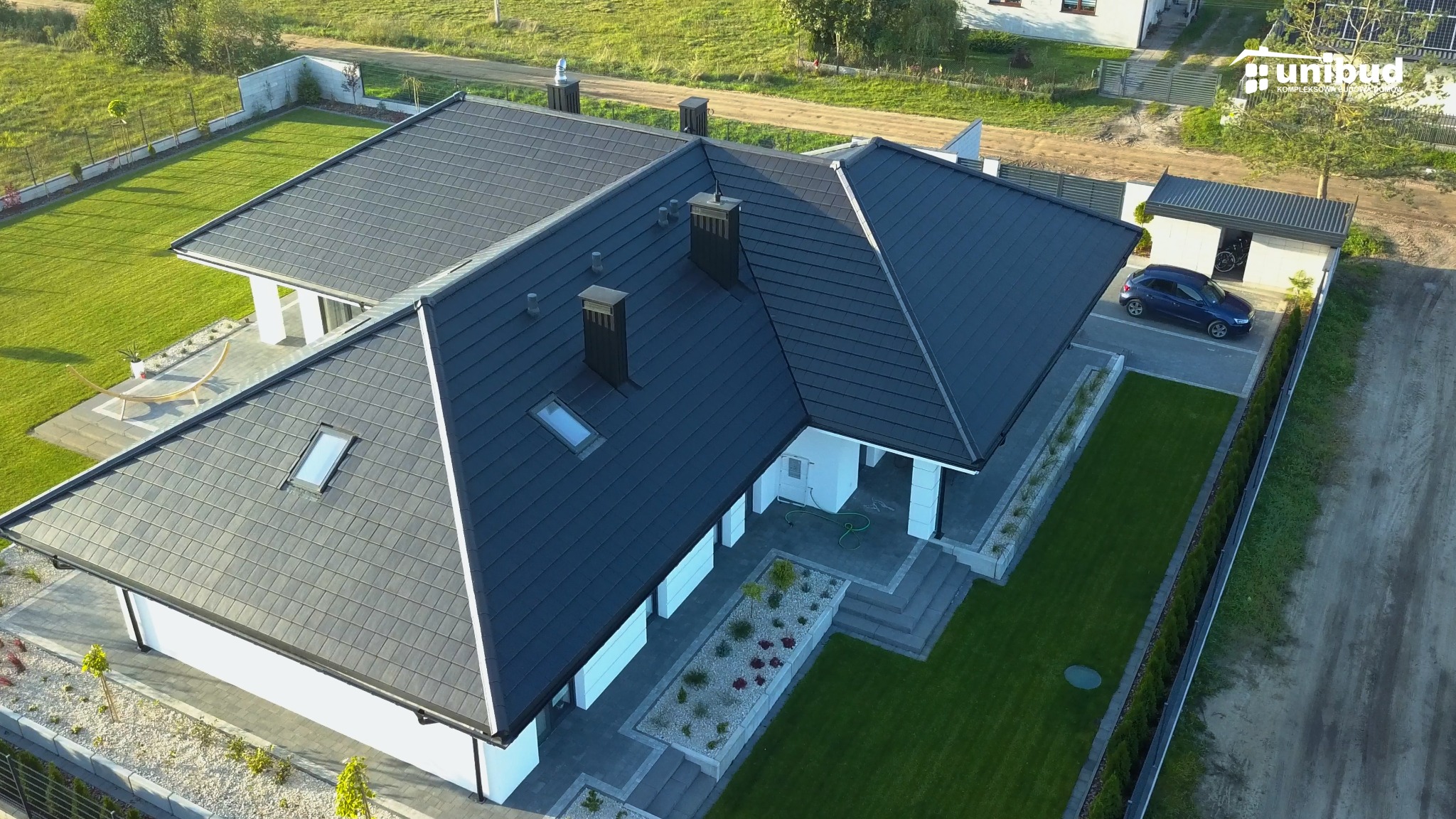 Widok z lotu ptaka na nowoczesny dom z ciemnym dachem, białymi ścianami i zadbanym trawnikiem, z widocznym garażem i samochodem osobowym.