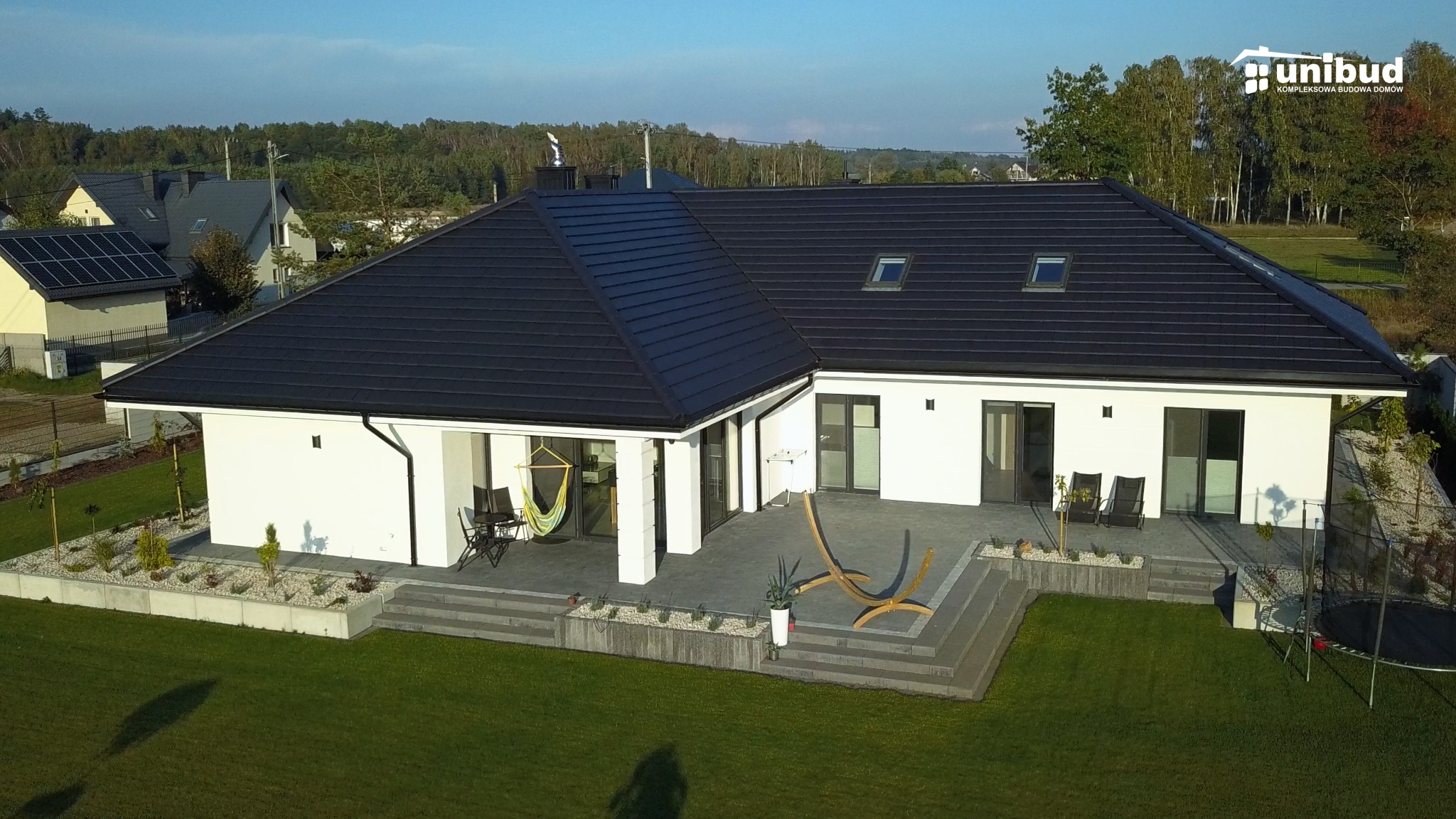 Nowoczesny dom murowany z czarnym dachem, białymi ścianami i przestronnym tarasem z drewnianymi leżakami i hamakiem, widok z lotu ptaka na tle zielonego ogrodu i lasu.